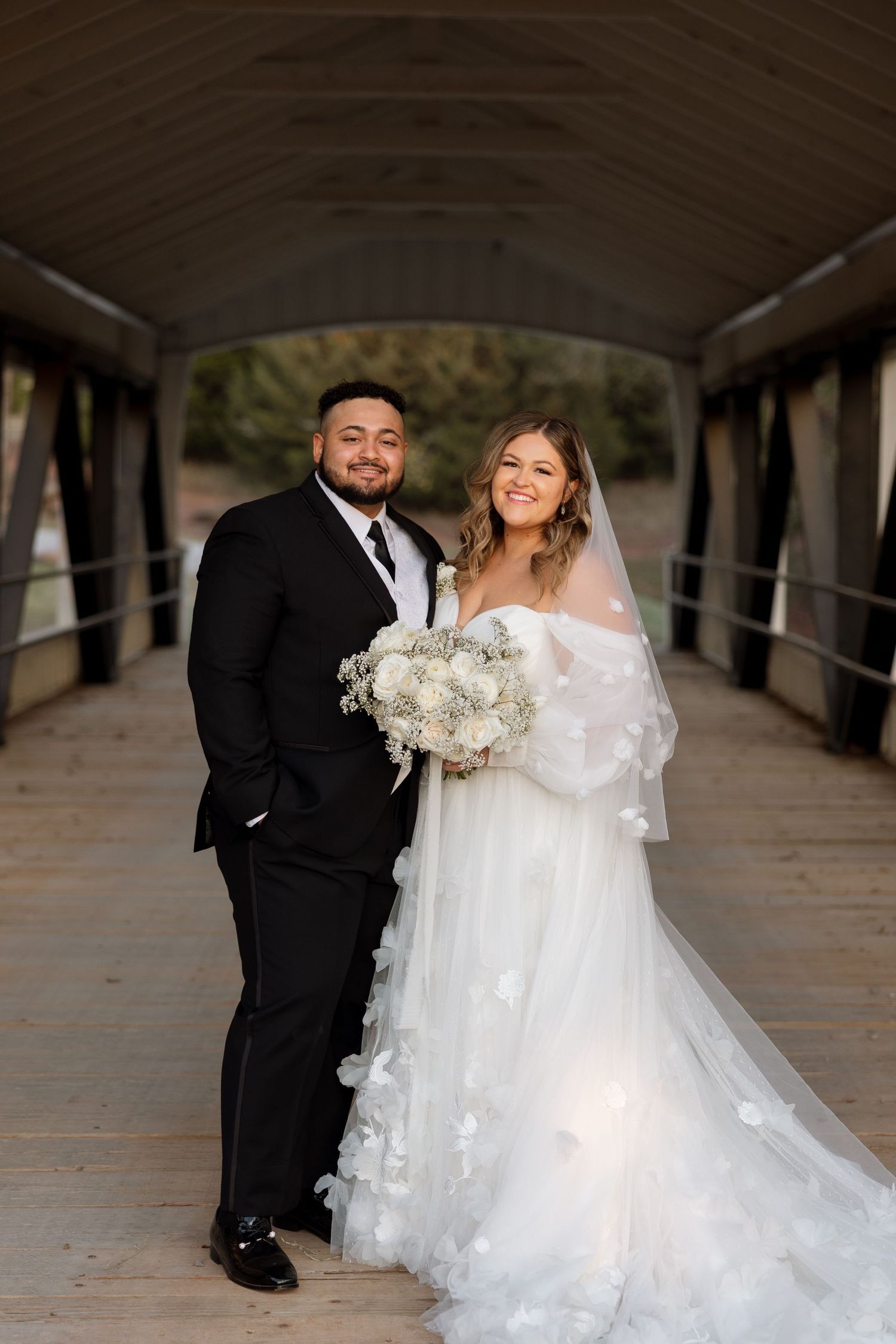 A joyful wedding portrait on a covered wooden bridge with white floral bouquet.