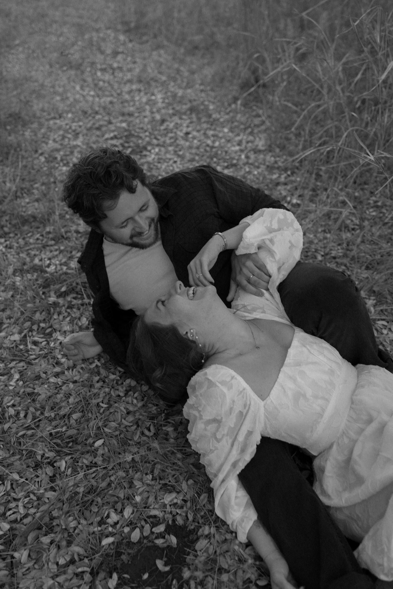 A black and white image of a couple laying together on fallen leaves in casual attire.