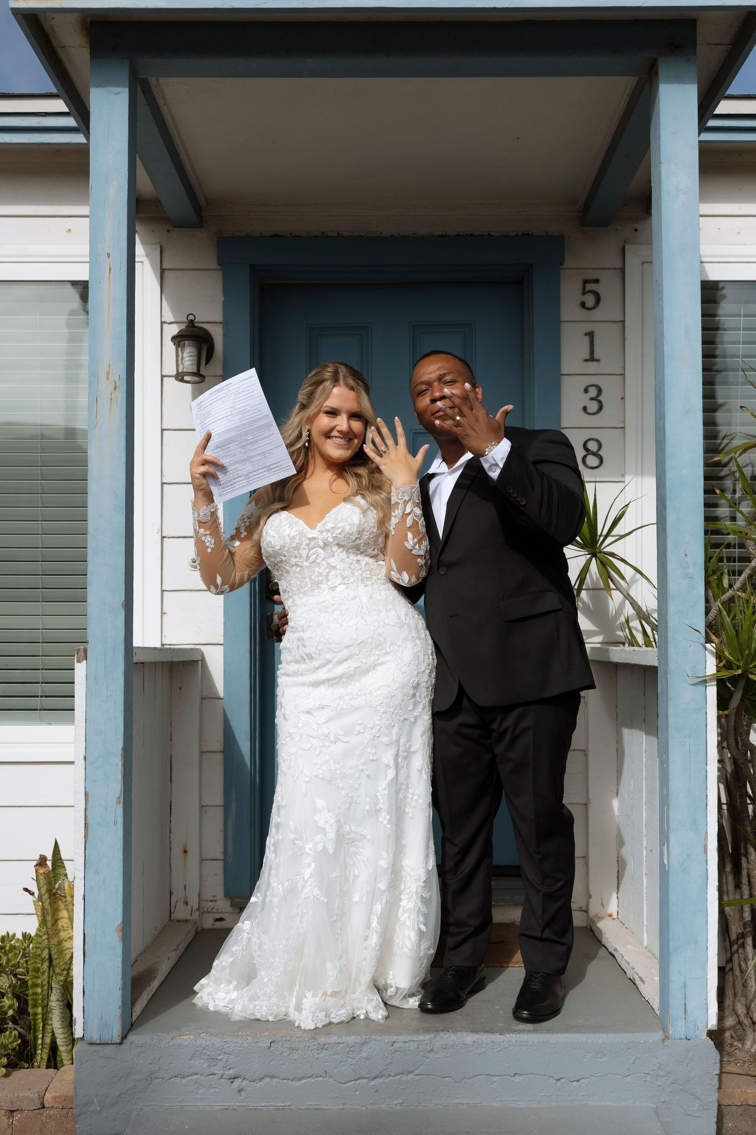 A wedding couple poses joyfully on a blue-trimmed front porch of a house with the number 5138.