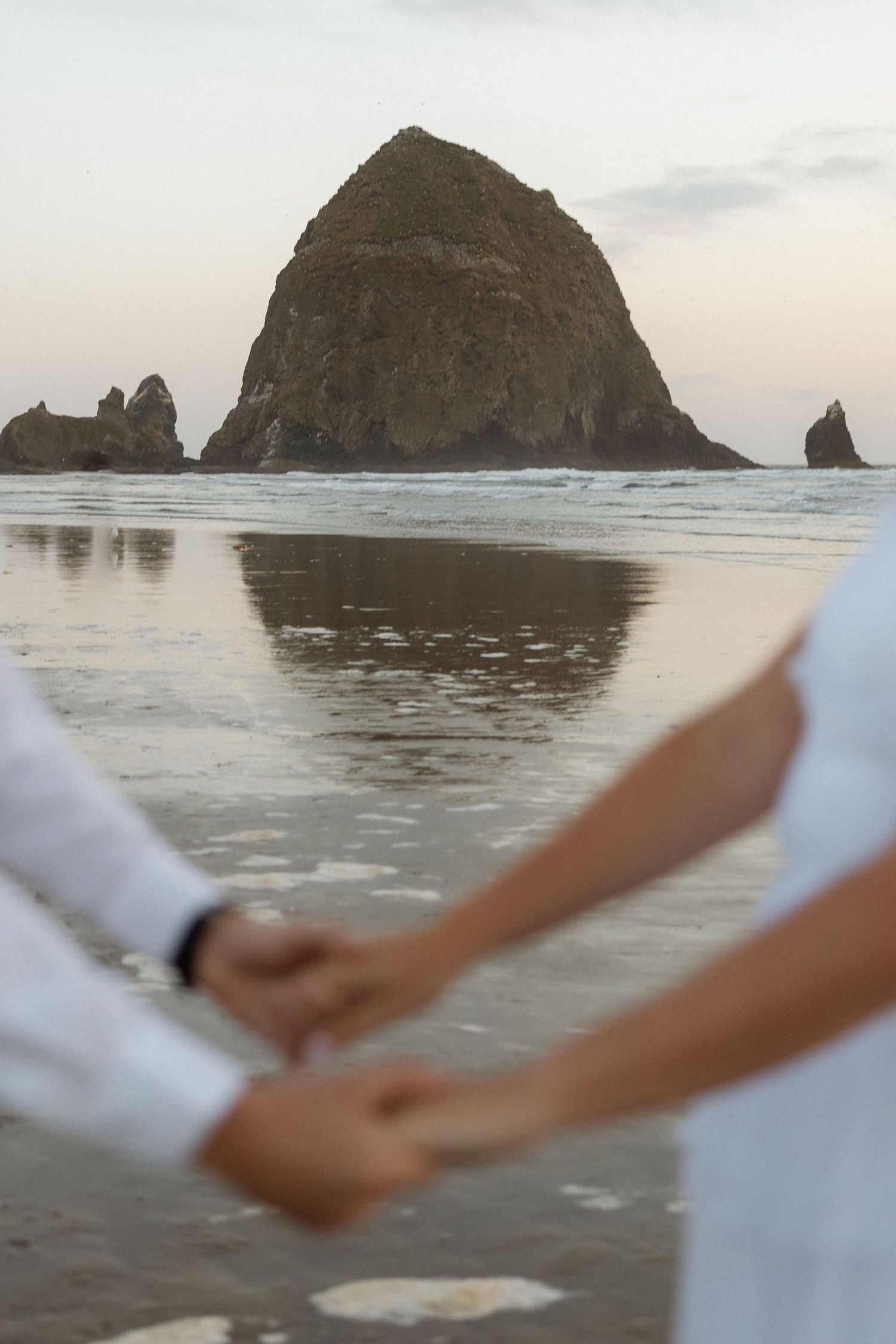 Couple holding hands on beach with iconic Haystack Rock at Cannon Beach, Oregon in background at sunset.