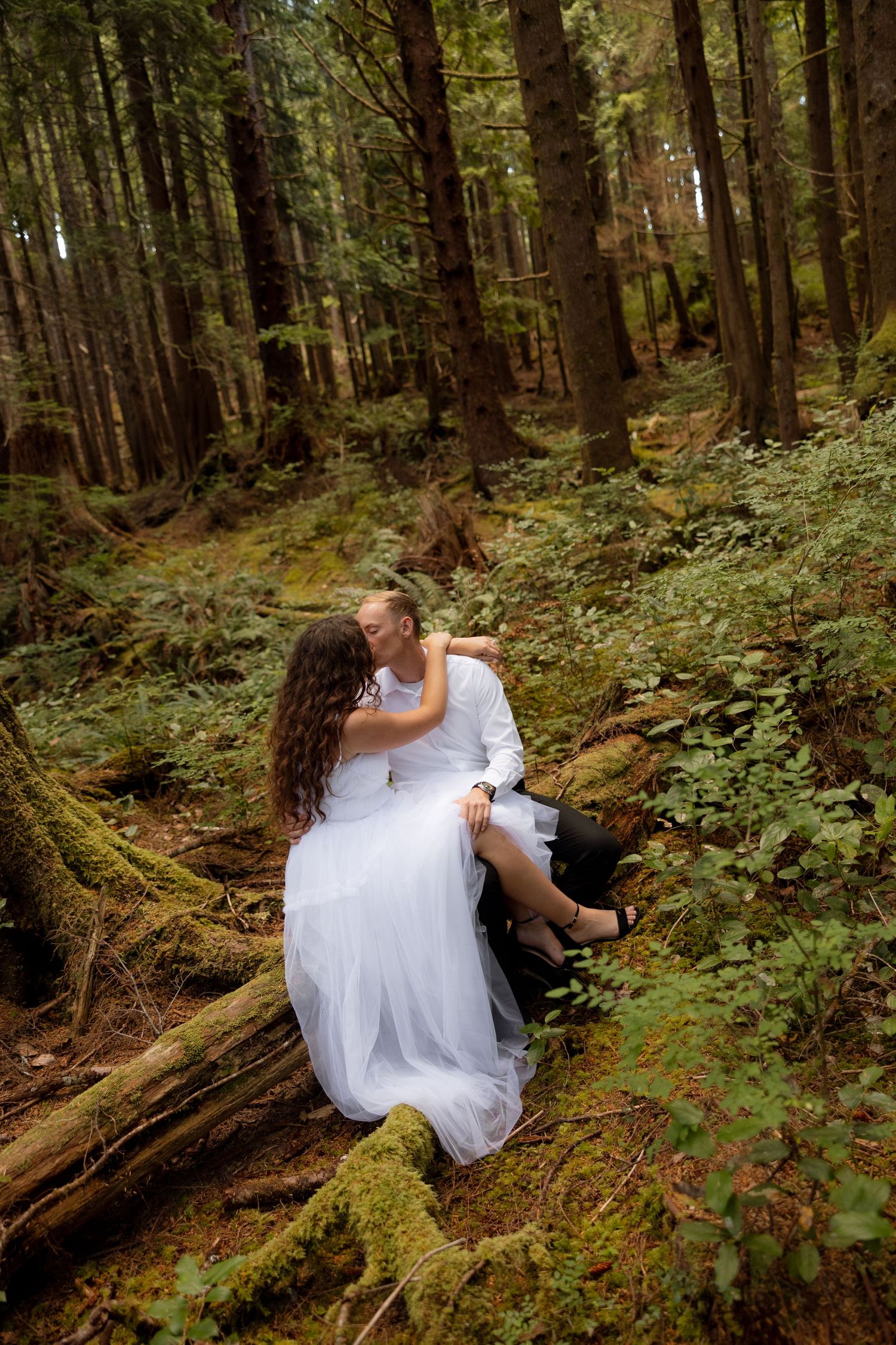 A romantic wedding pose on a moss-covered log in a dense forest setting.
