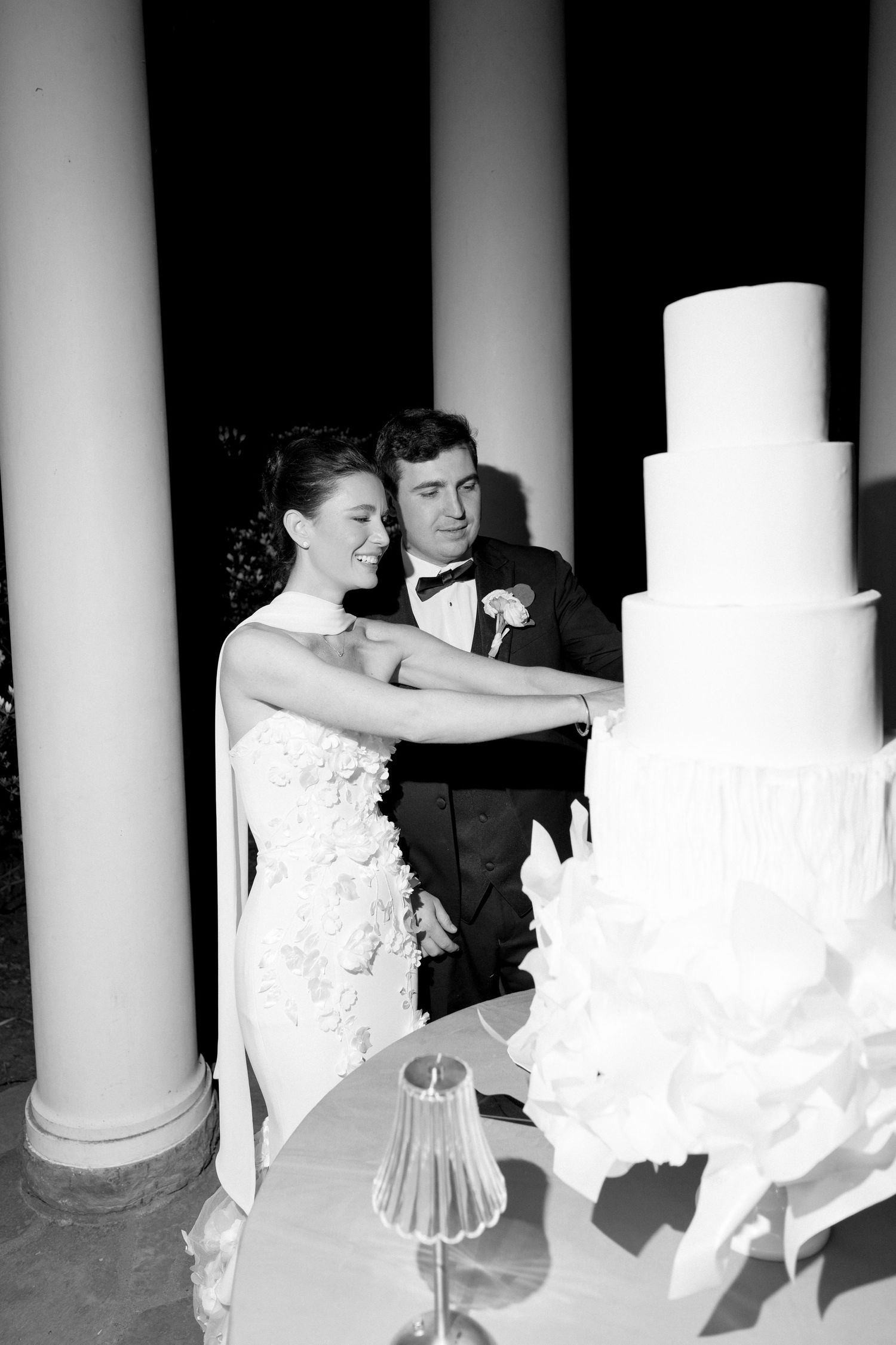 Bride and groom cut elegant five-tier white wedding cake beside grand columns in black and white photo.