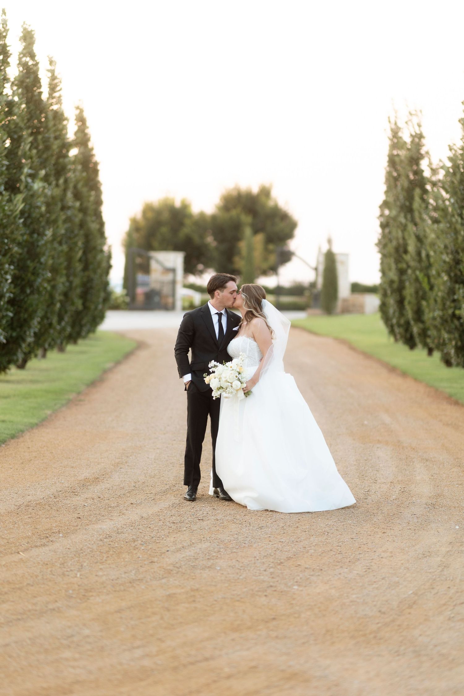 A couple in wedding attire poses romantically on a tree-lined dirt path during golden hour.
