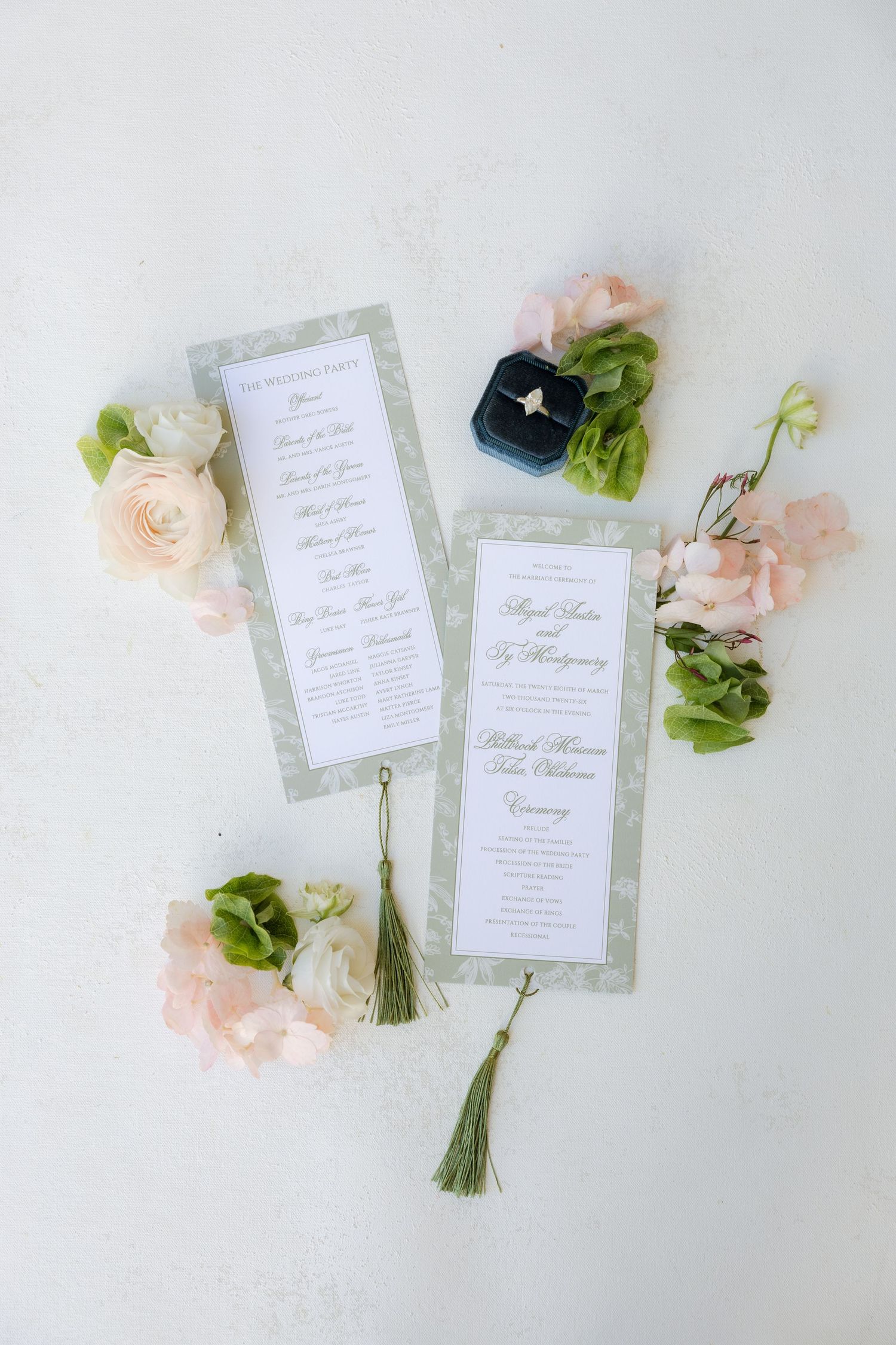 Elegant sage green wedding ceremony programs with gold tassels displayed with pink flowers and ring box.