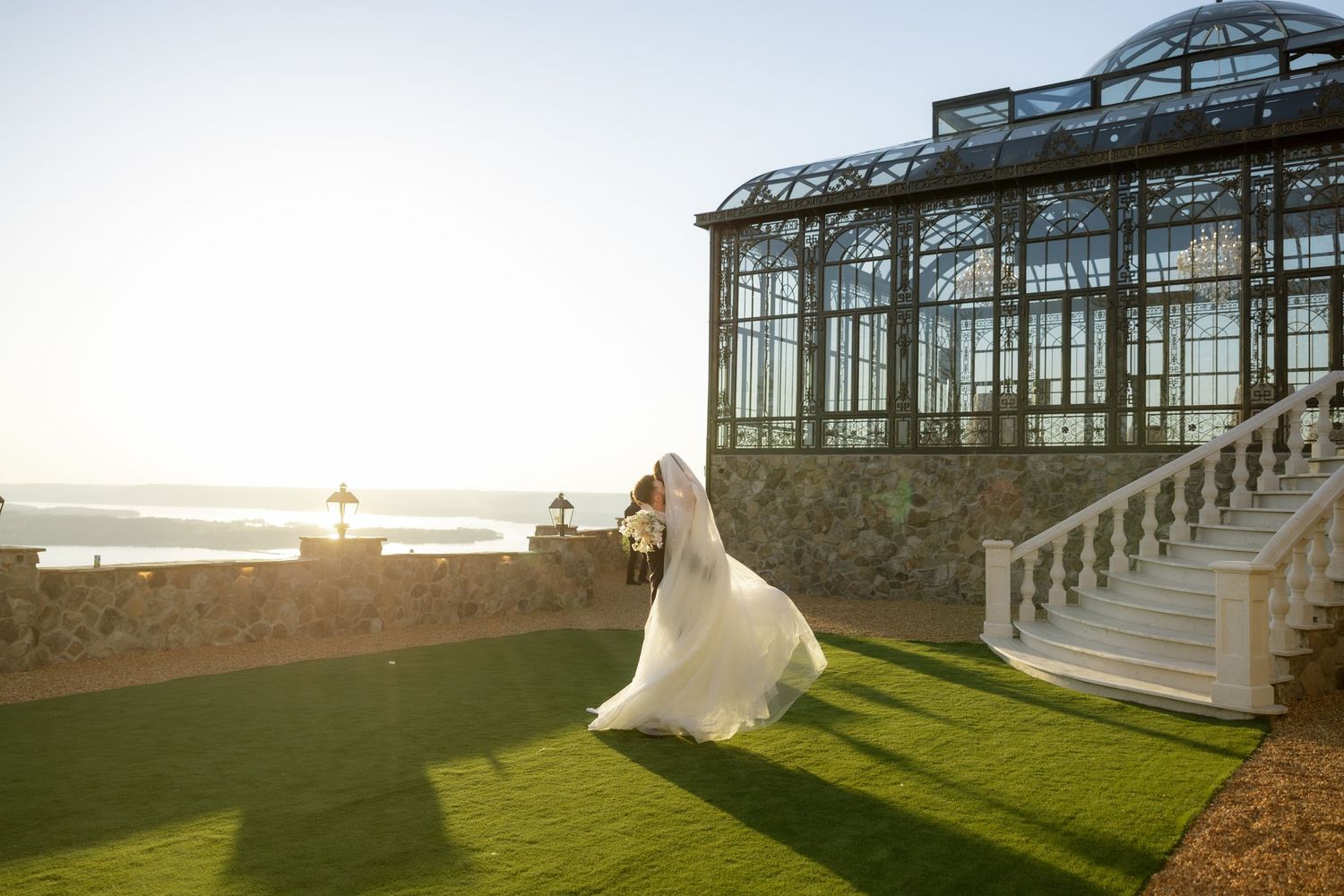 Bride in flowing white gown walks on green lawn beside elegant glass greenhouse venue with ocean views.