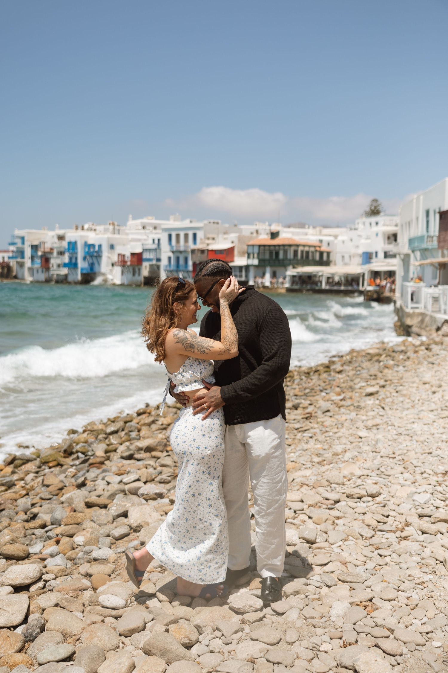 A couple shares a romantic moment on a rocky beach with traditional Greek waterfront buildings behind them.