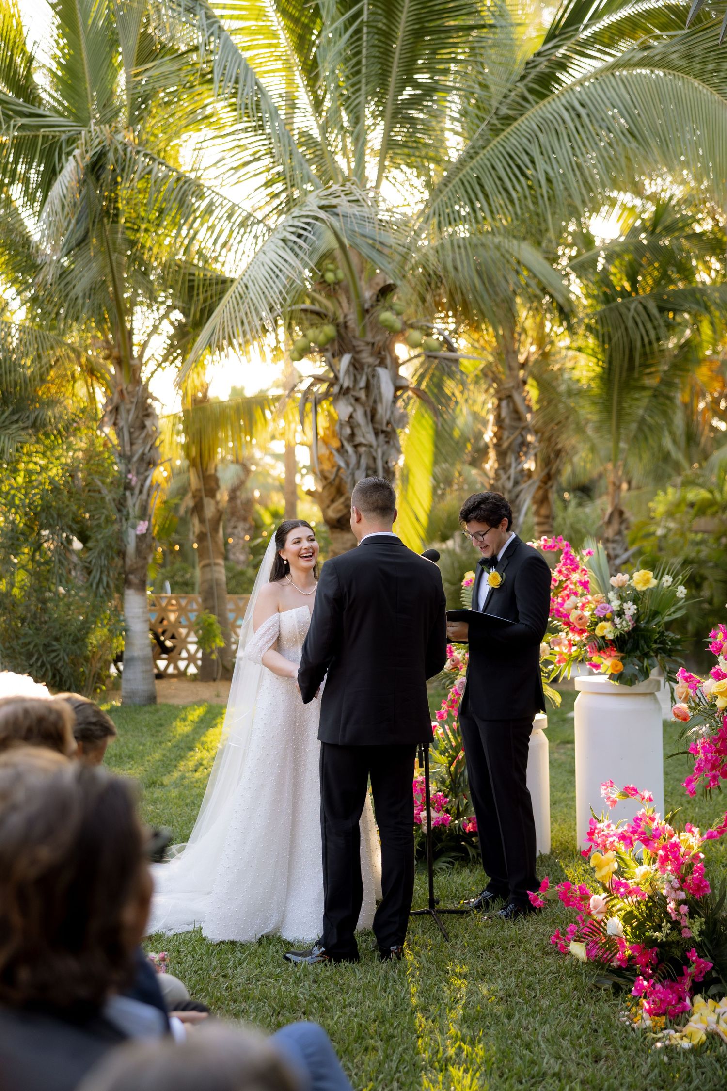 Bride and groom exchange vows at an outdoor ceremony surrounded by palm trees and vibrant pink floral arrangements.