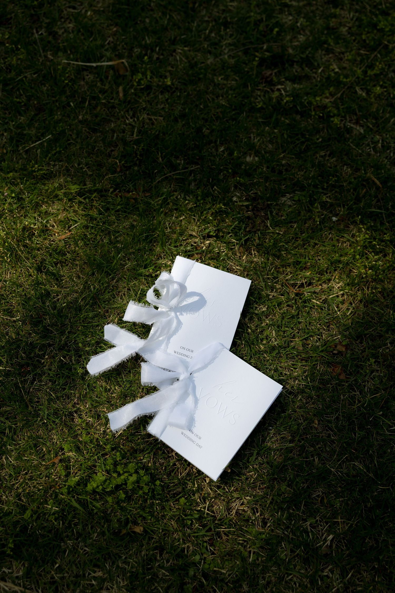 Two white wedding vow books tied with ribbon bows resting on green grass.