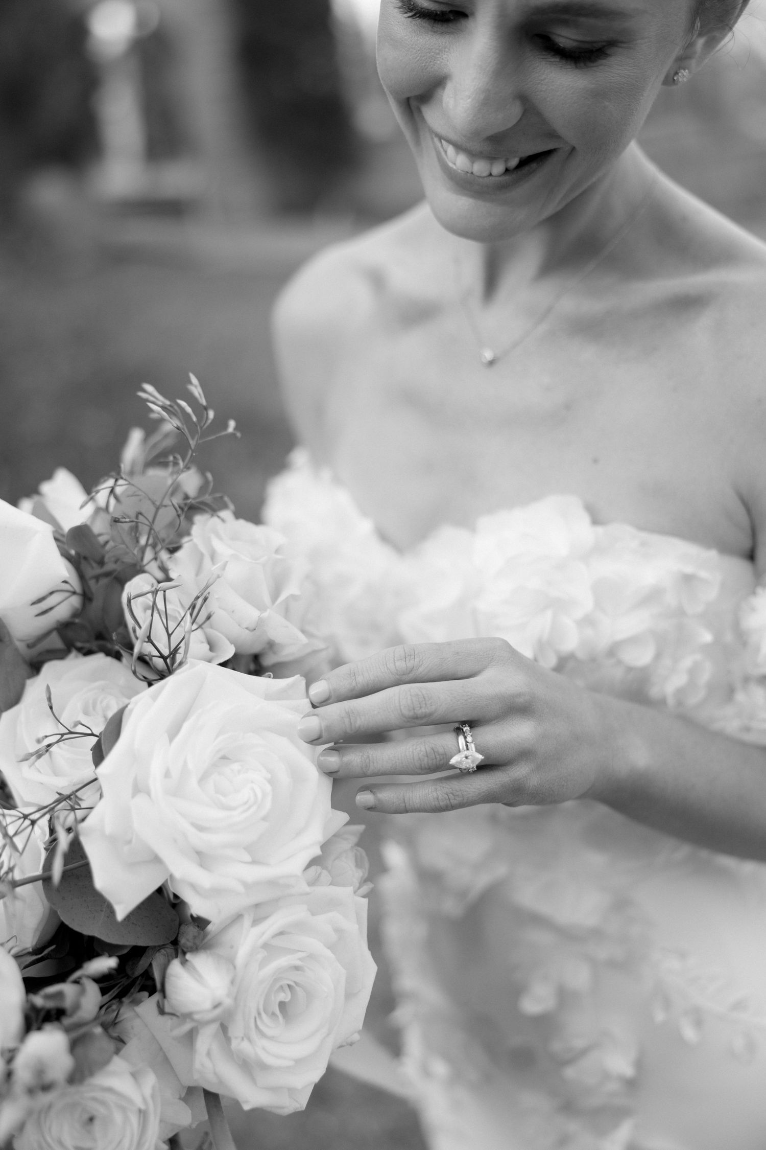 Smiling bride in strapless gown holds white rose bouquet displaying diamond engagement ring in black and white photo.