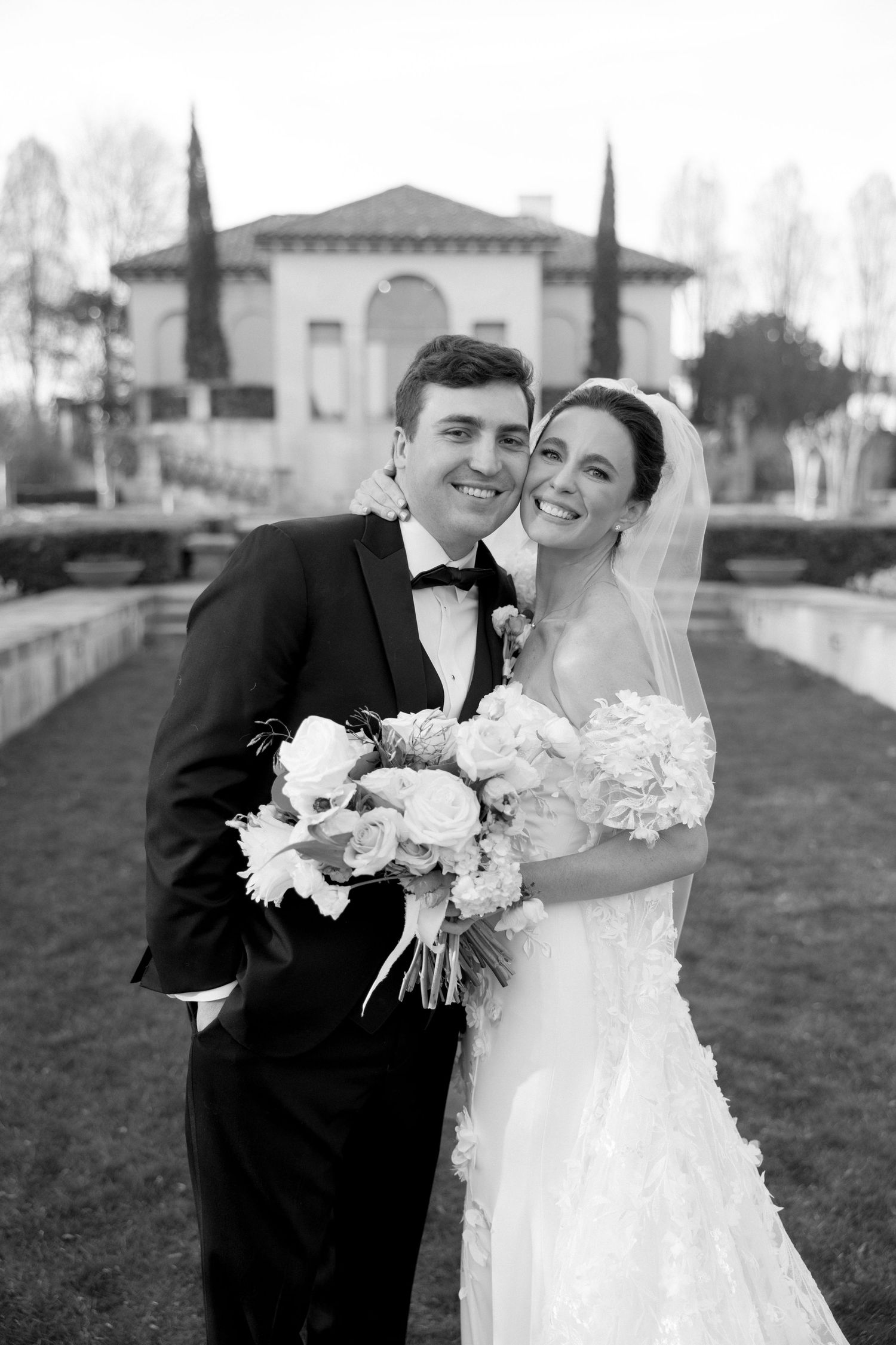 Black and white portrait of smiling newlyweds holding bouquet outside Italian-inspired wedding venue.