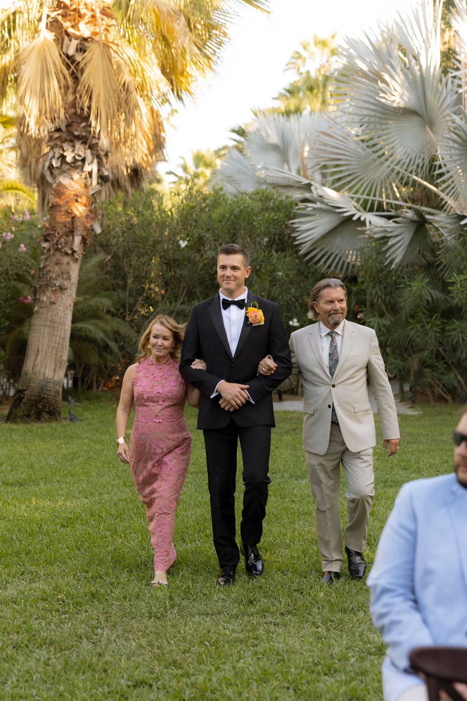 Groom in black tuxedo walks with parents across green lawn at outdoor wedding ceremony.
