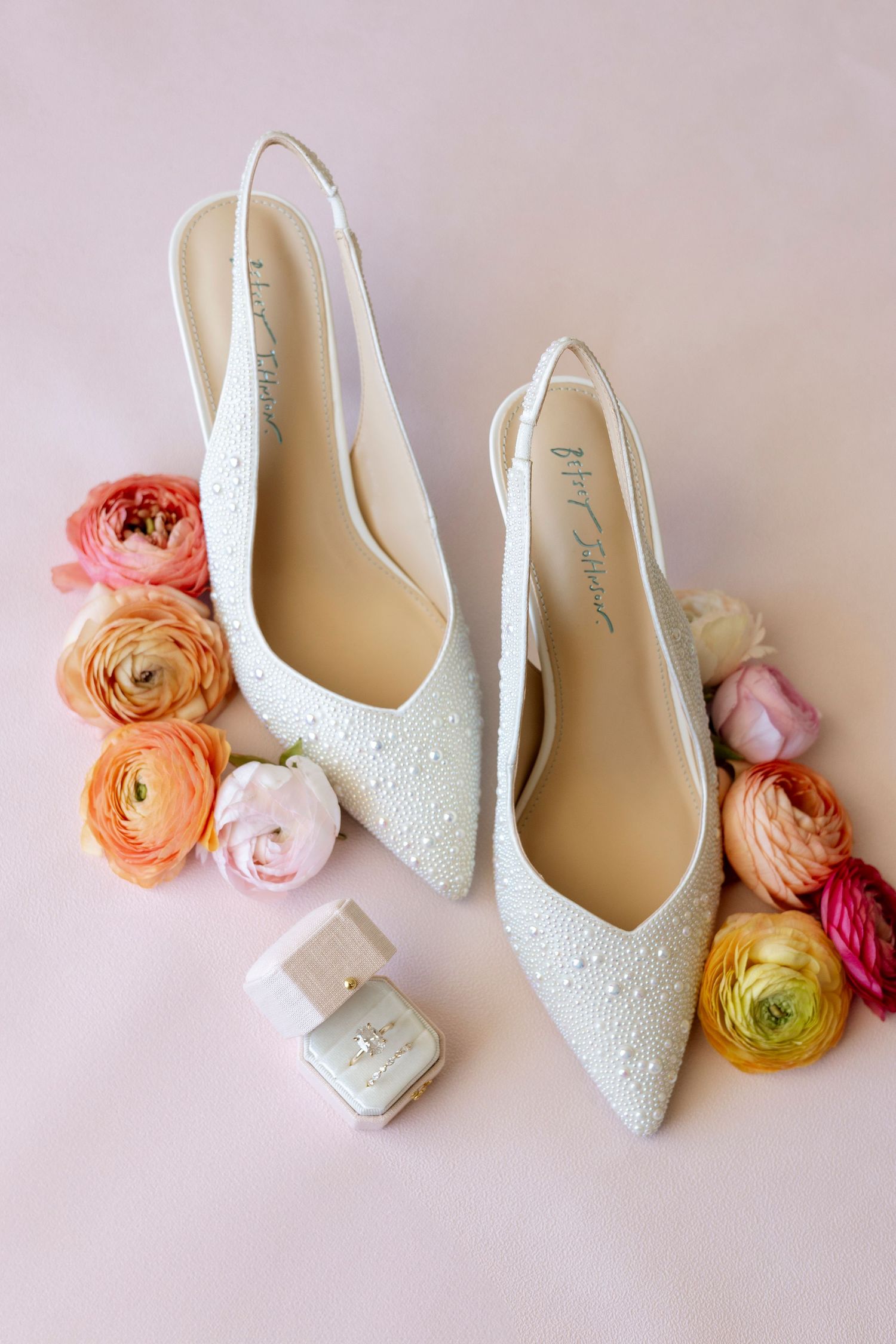 White bridal shoes decorated with delicate beading displayed alongside pink and peach ranunculus flowers.