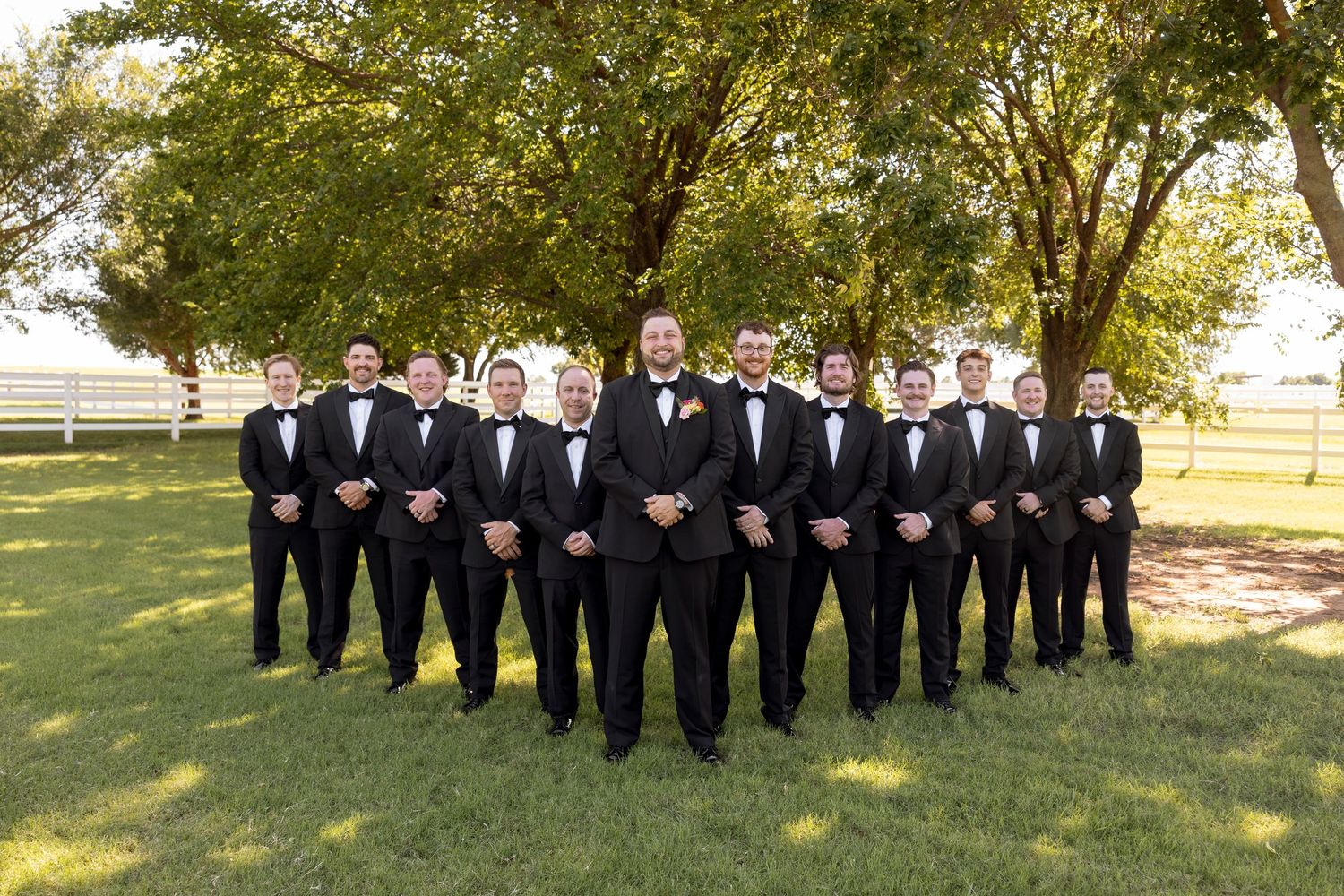 A row of people in formal black suits and ties stand together on grass in front of trees for a group photo.