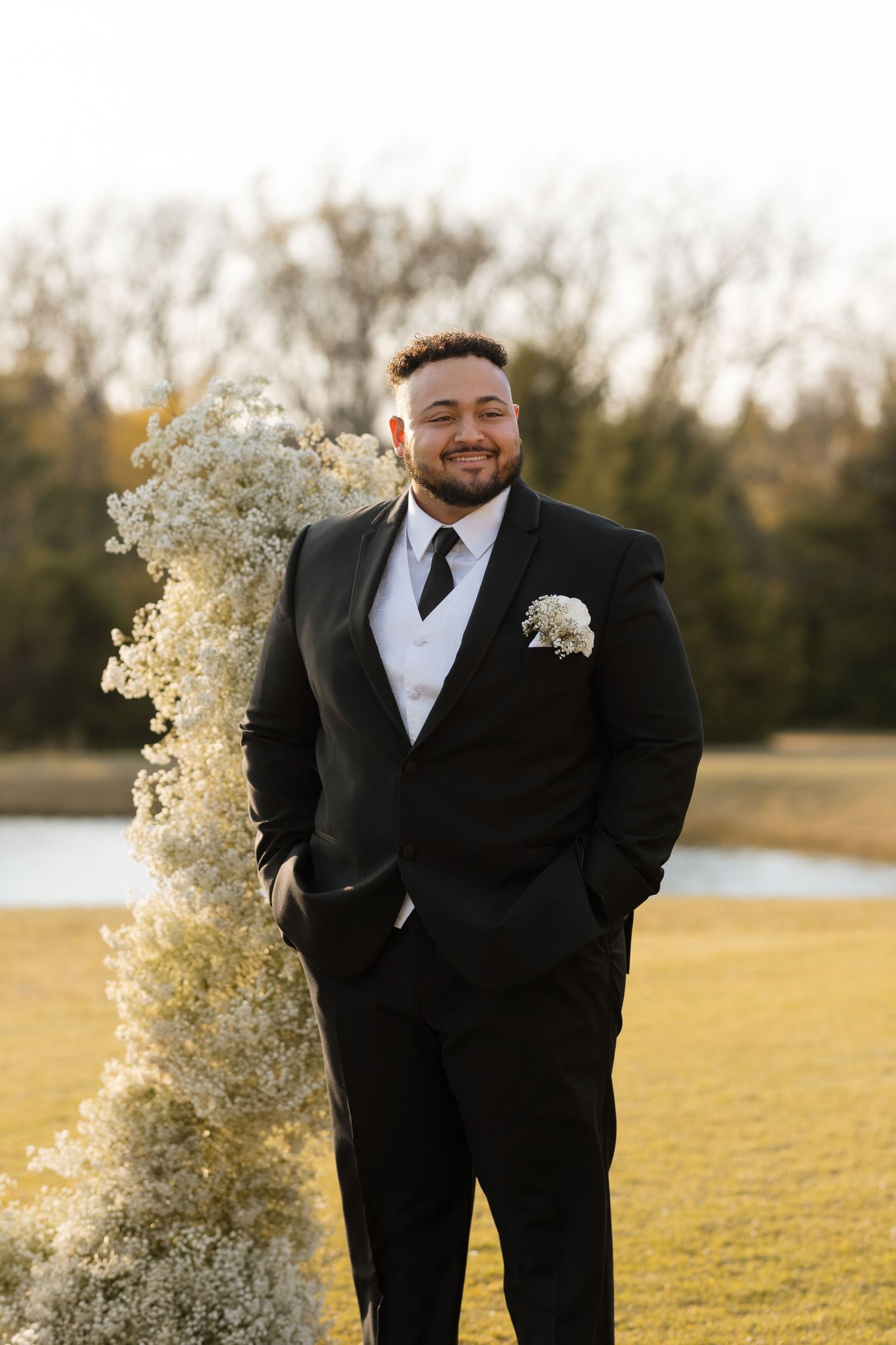 Wedding party member in black tuxedo stands by decorative floral arrangement outdoors.