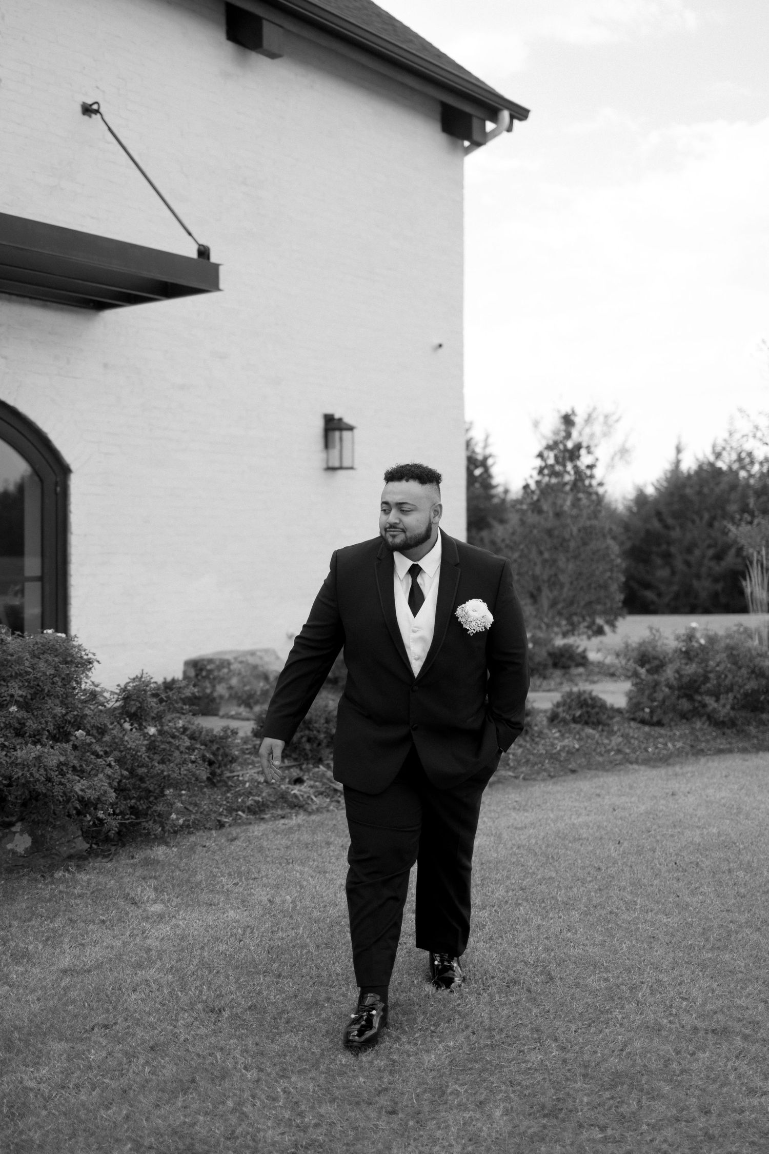 A groom in a black tuxedo walks outside a white building on his wedding day.