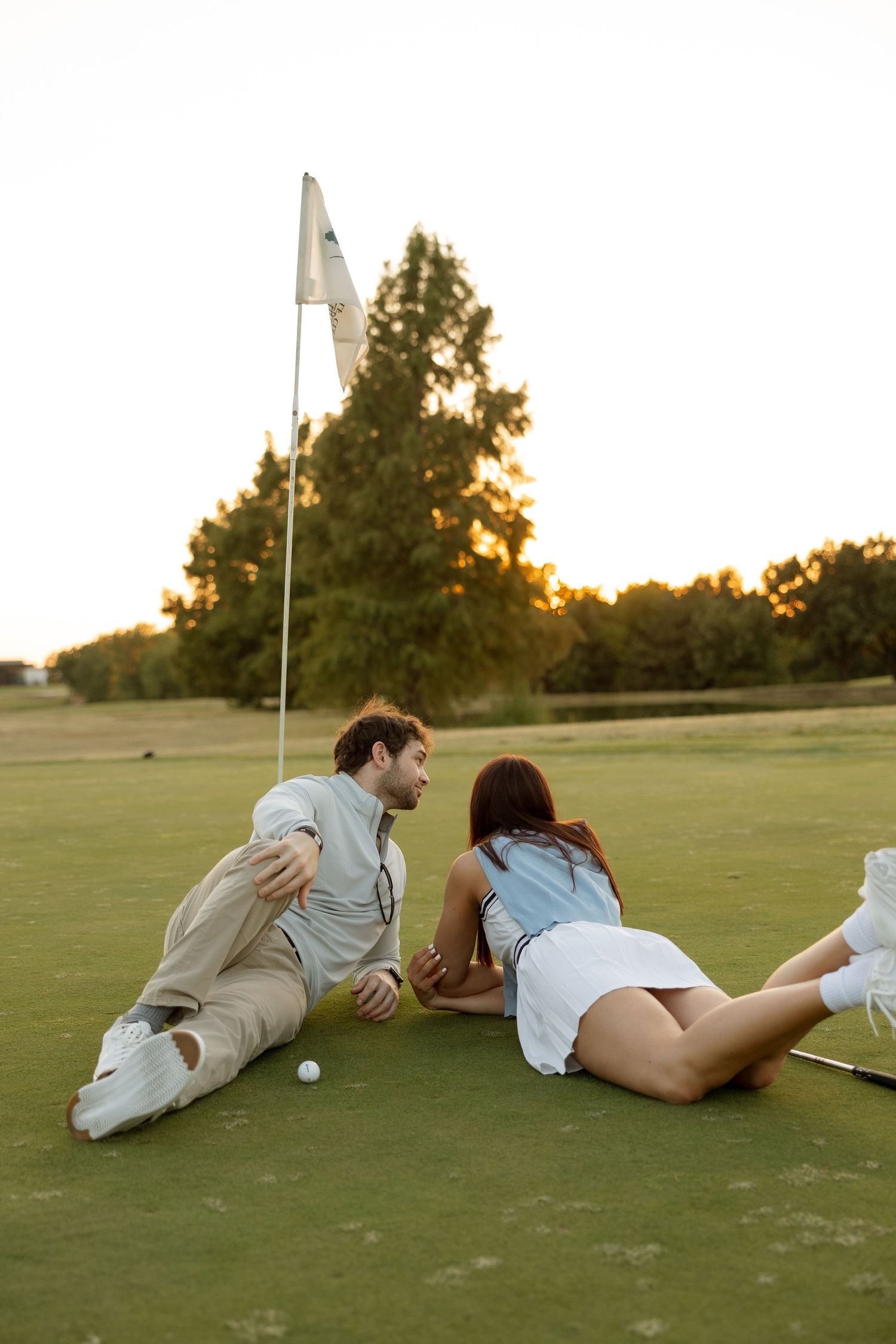 A couple lounges on a golf green at sunset near a flag pole having a romantic moment.