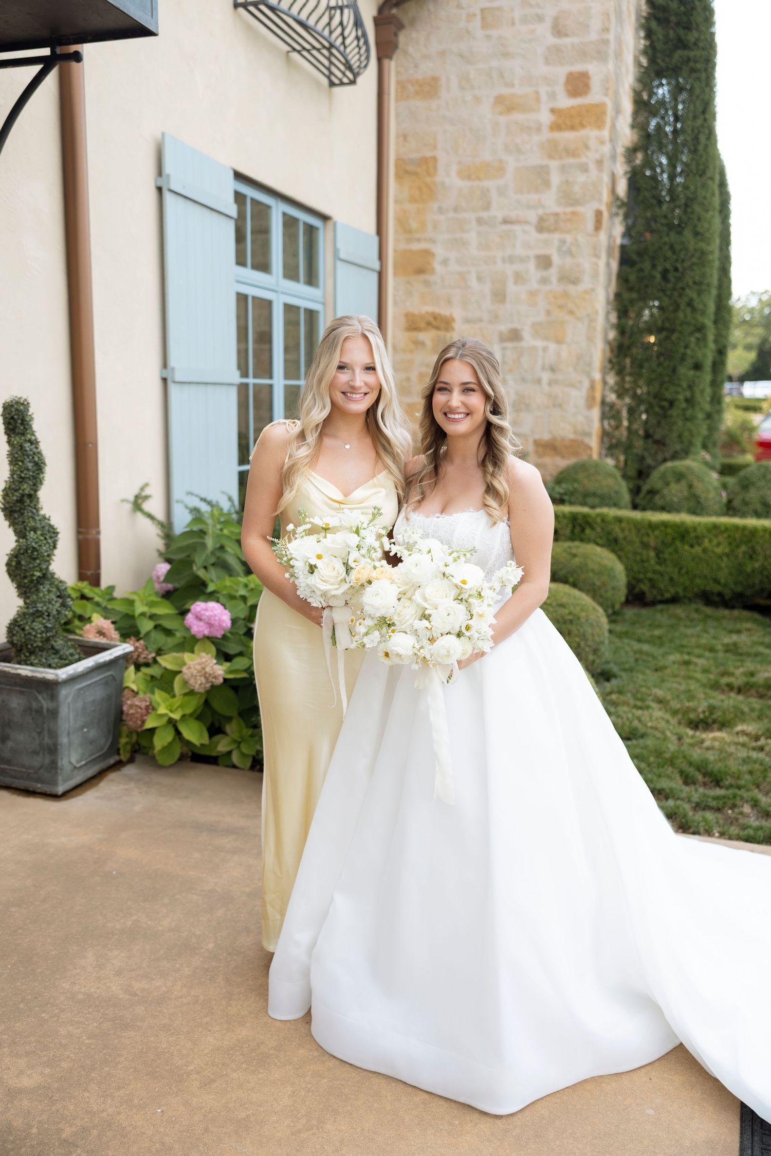 Two women in formal attire stand together holding white floral bouquets against a stone building with blue shutters.