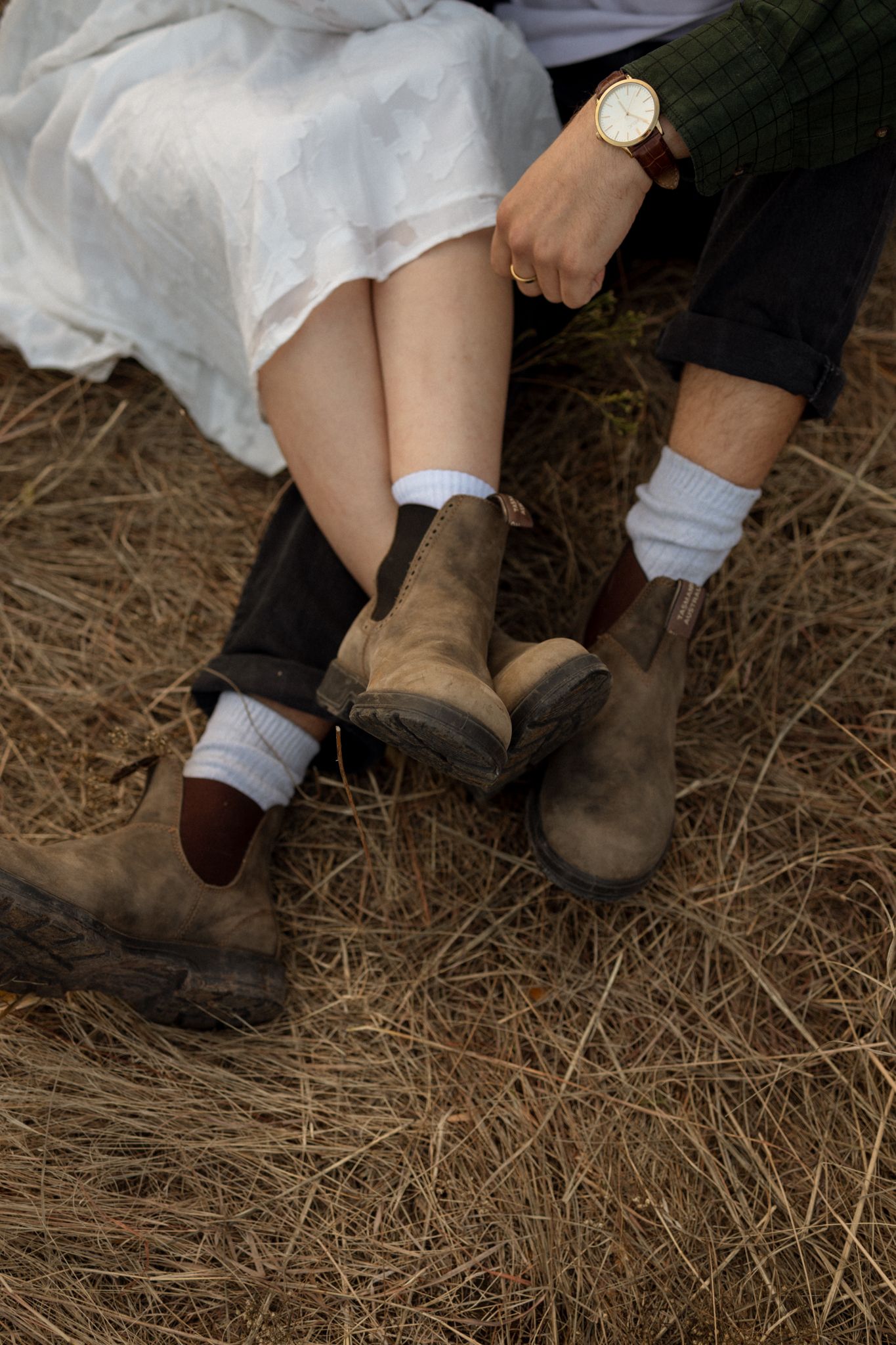 Two pairs of boots with white socks shown against dried grass, one in a white dress and dark pants.