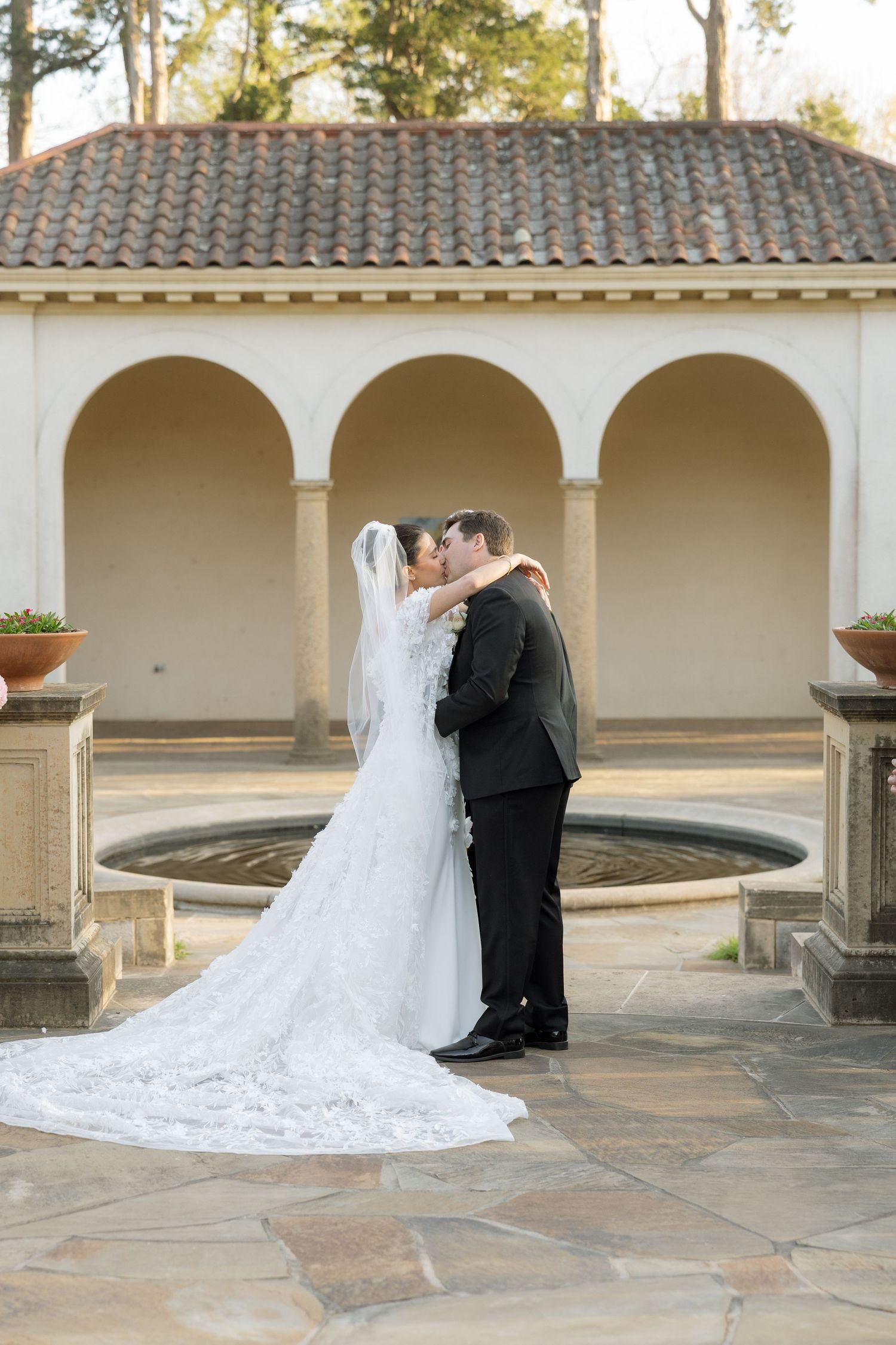 Bride and groom share first kiss in front of elegant arched stone fountain at outdoor wedding venue.
