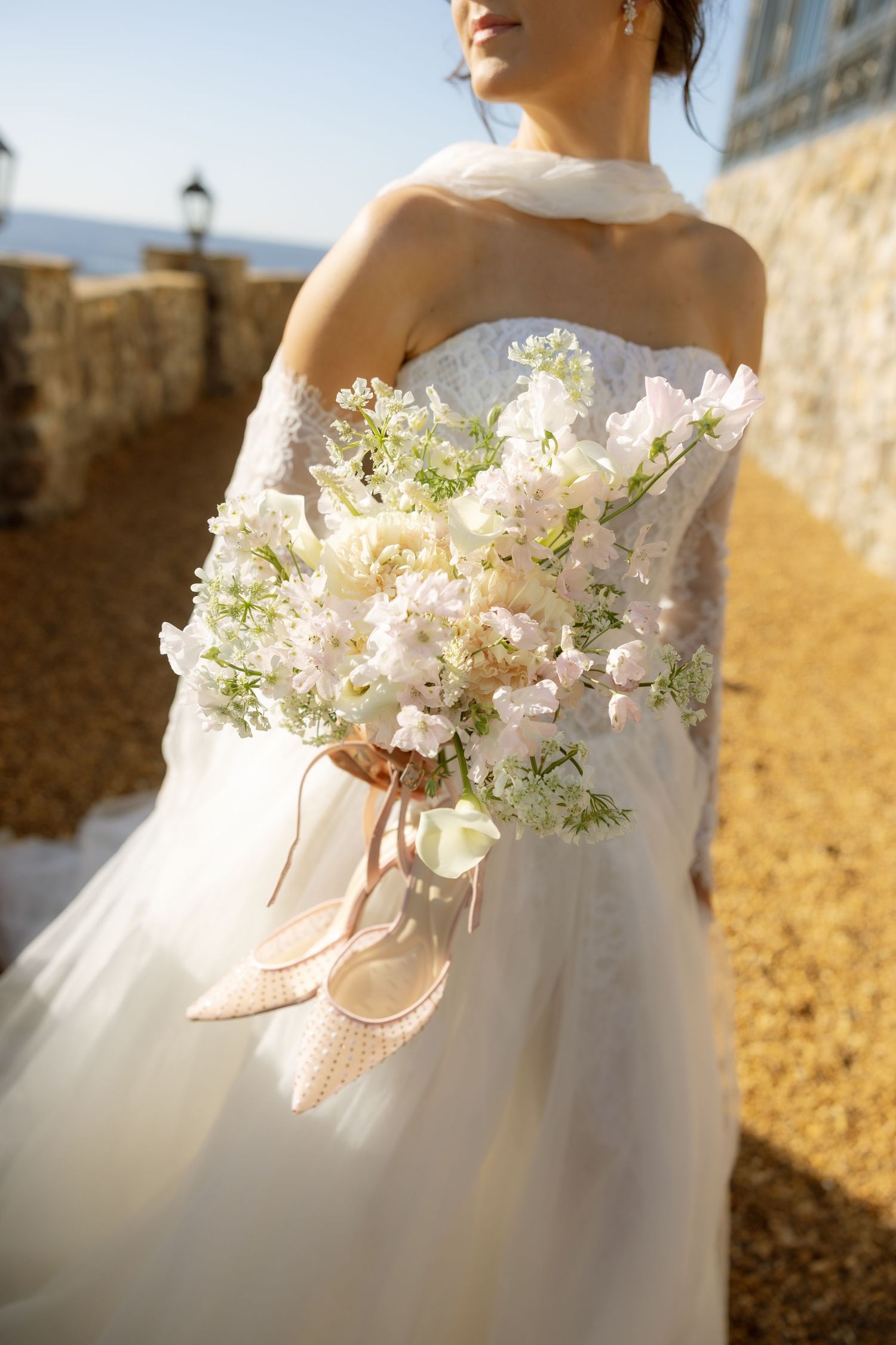 Bride in strapless white gown holding elegant bouquet of white flowers with blush ribbons and heels outdoors.