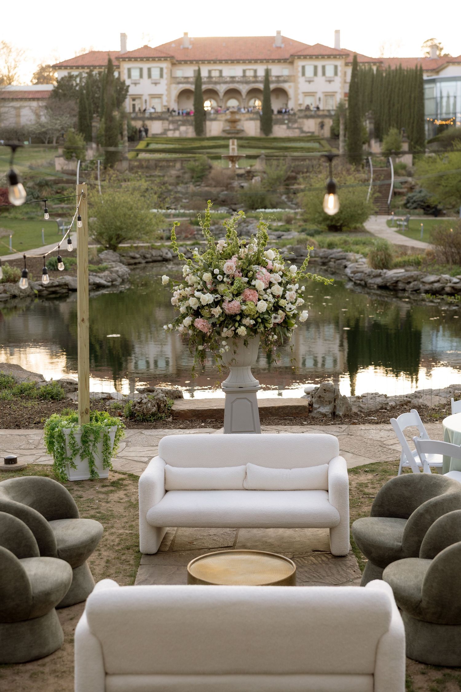 Elegant outdoor wedding lounge with white sofas, floral arrangement, and pond reflecting mansion.