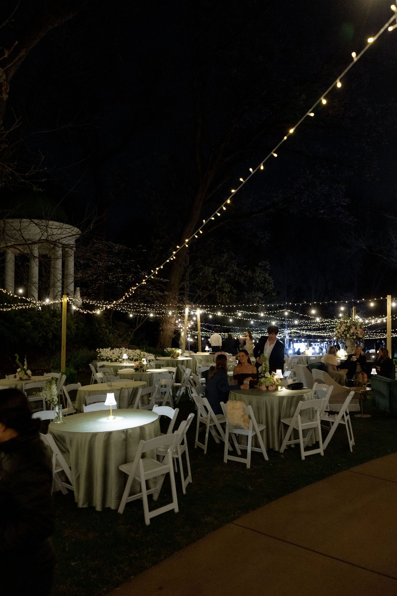 Outdoor wedding reception at night with string lights, round tables, white chairs, and glowing centerpieces.