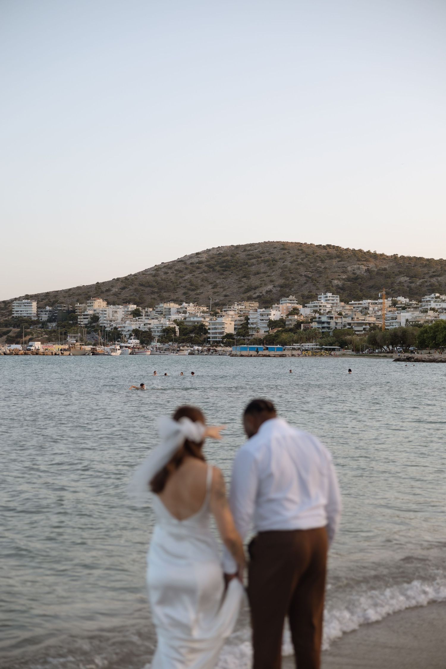 A couple stands at the water's edge during sunset with a coastal cityscape and hillside in the background.