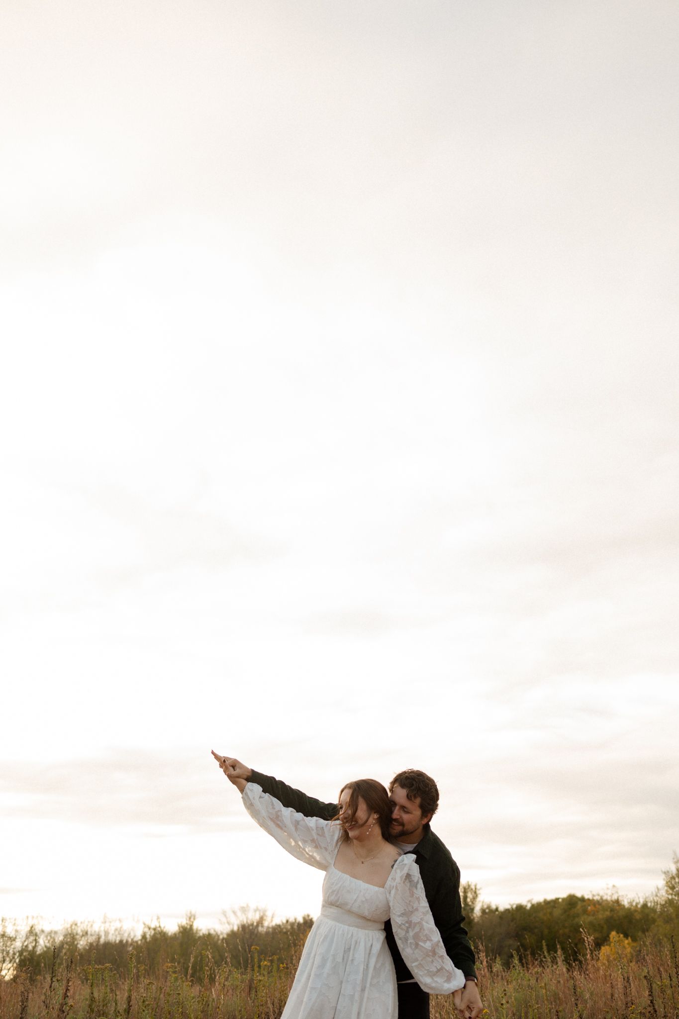 A romantic couple stands in a golden field at sunset, with one pointing toward the horizon.