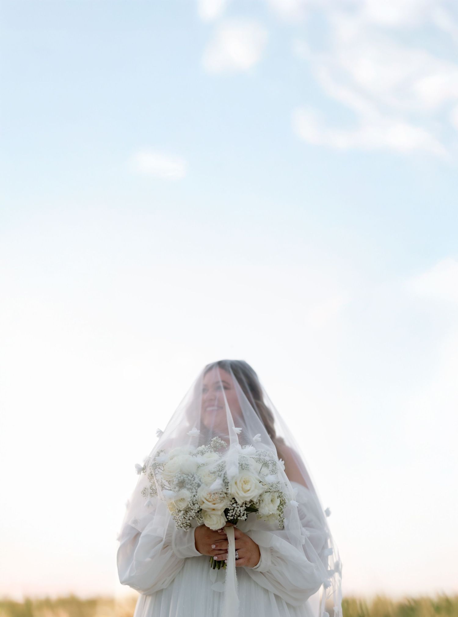 A bride holds a white bouquet against a bright sky while wearing a delicate veil.