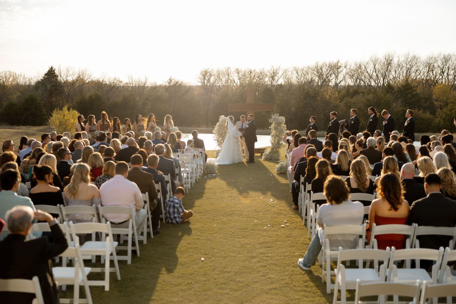 Sunset wedding ceremony with guests seated in white chairs on grassy outdoor venue.