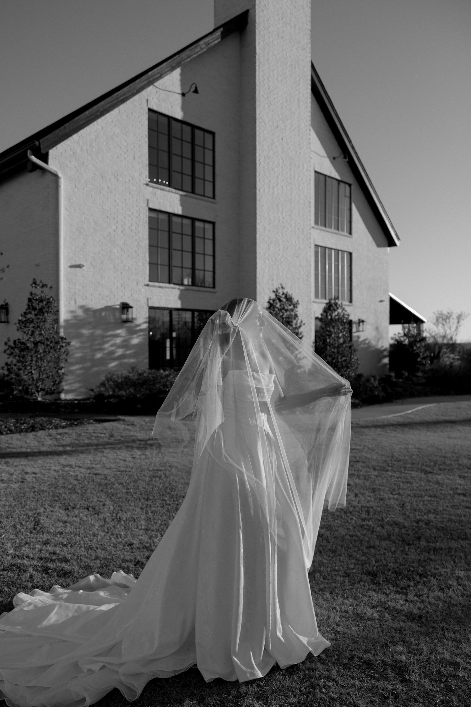 A ghostly figure draped in a flowing white wedding gown stands in front of a modern white house in black and white.