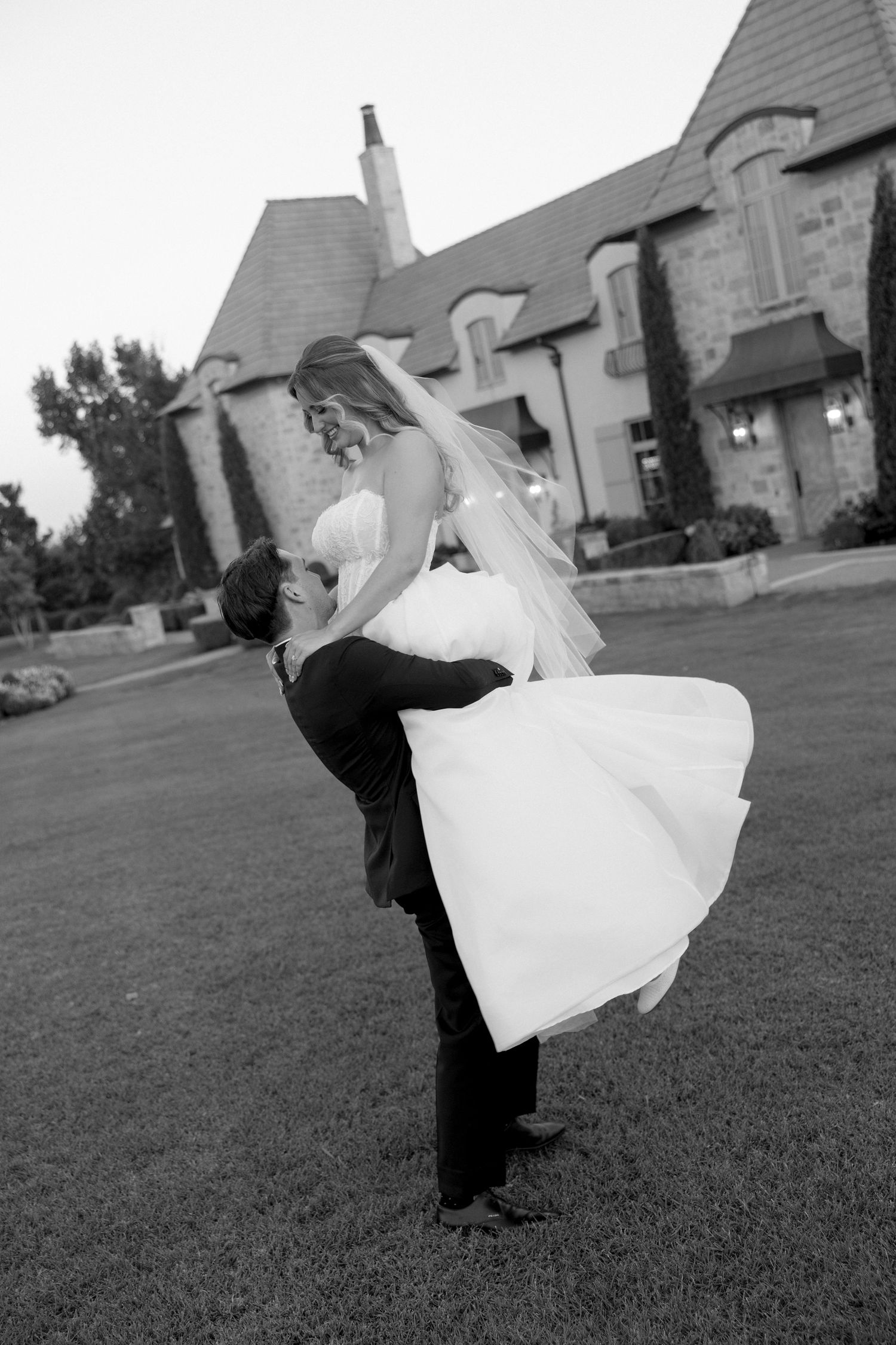 A romantic black and white wedding photo showing a couple embracing on a lawn with a stately building in the background.