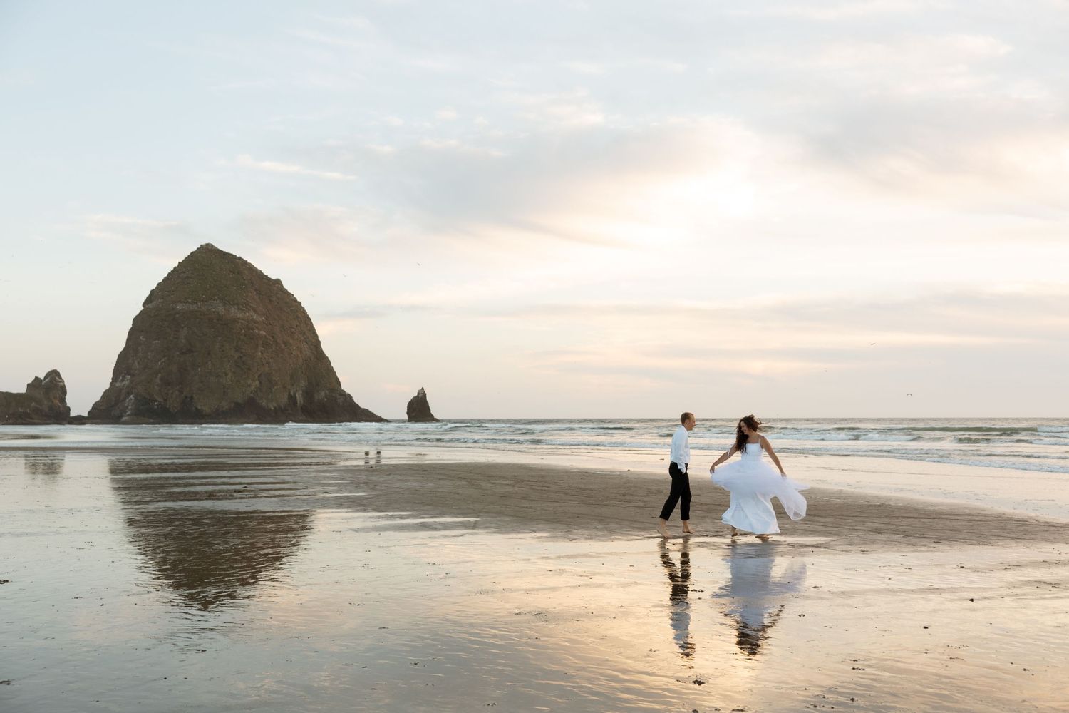 A couple walks along Cannon Beach at sunset with Haystack Rock in the background.