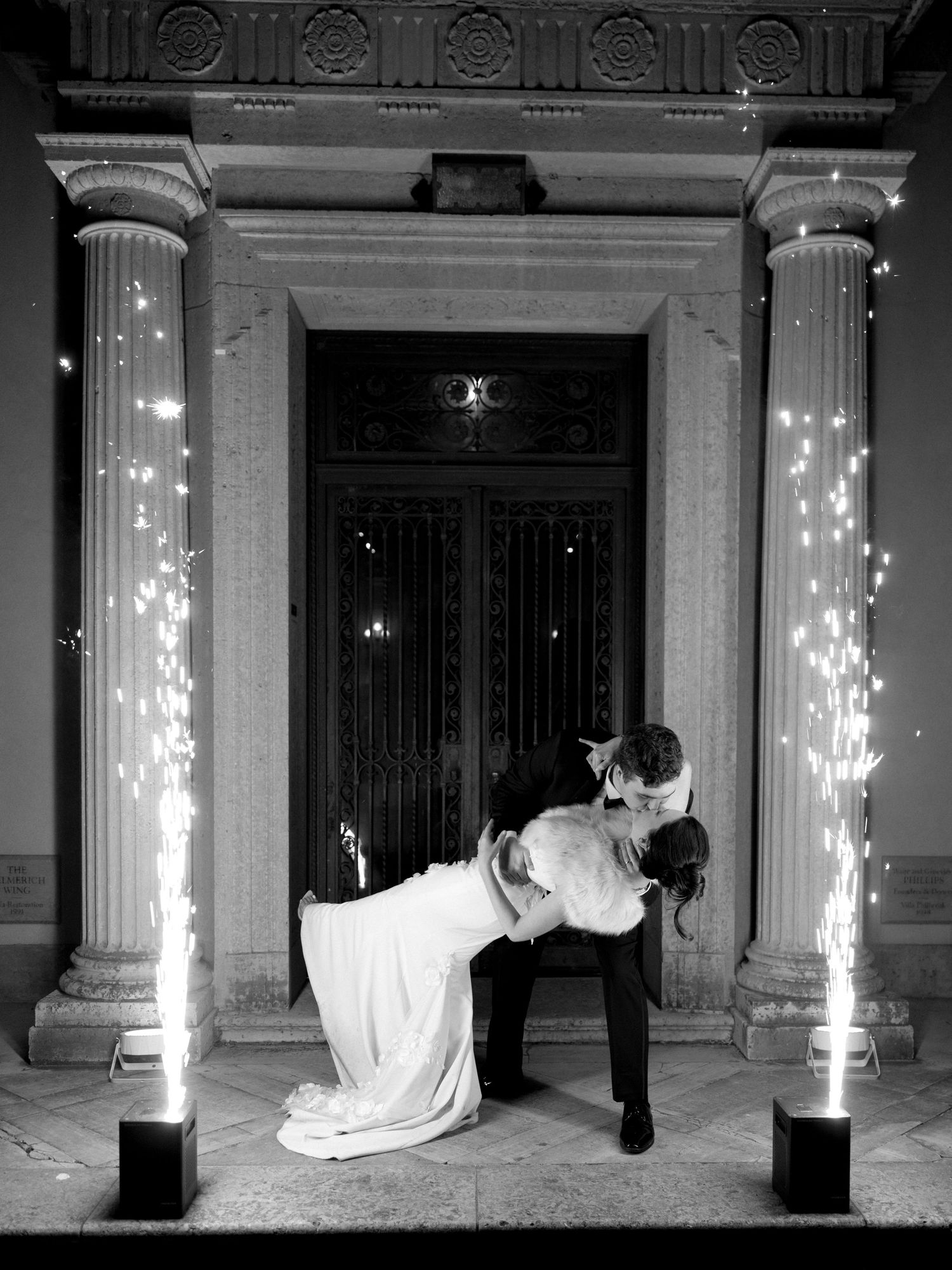 Black and white photo of bride and groom kissing between sparkling fountain fireworks at elegant venue entrance.