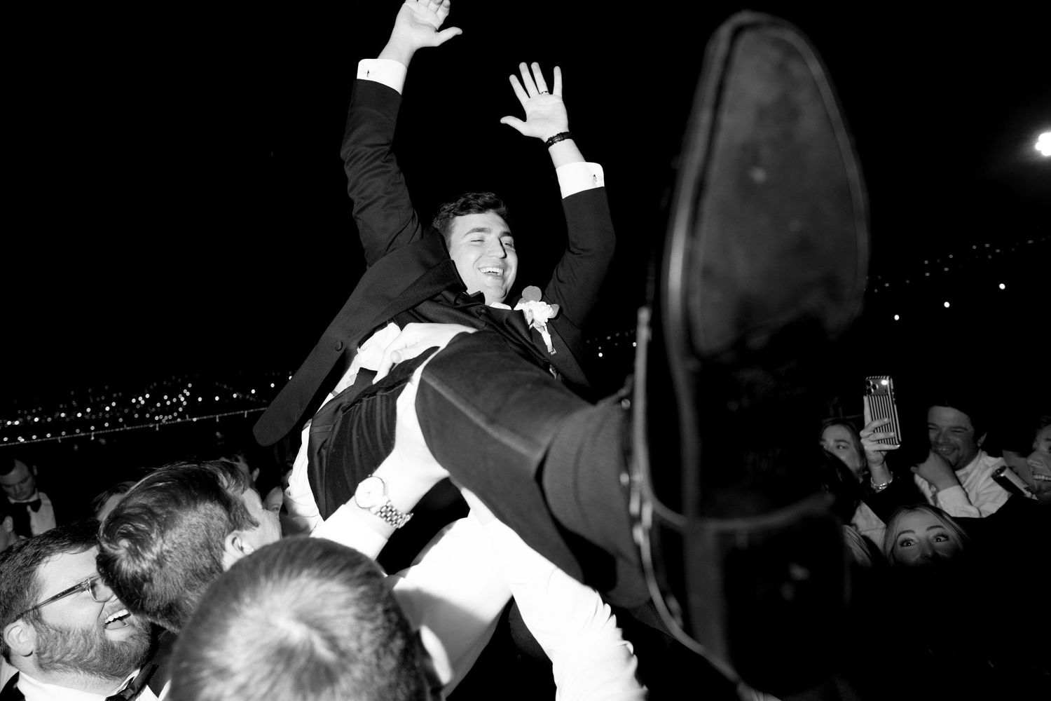 Groom being lifted by crowd during lively wedding reception celebration in black and white photography.