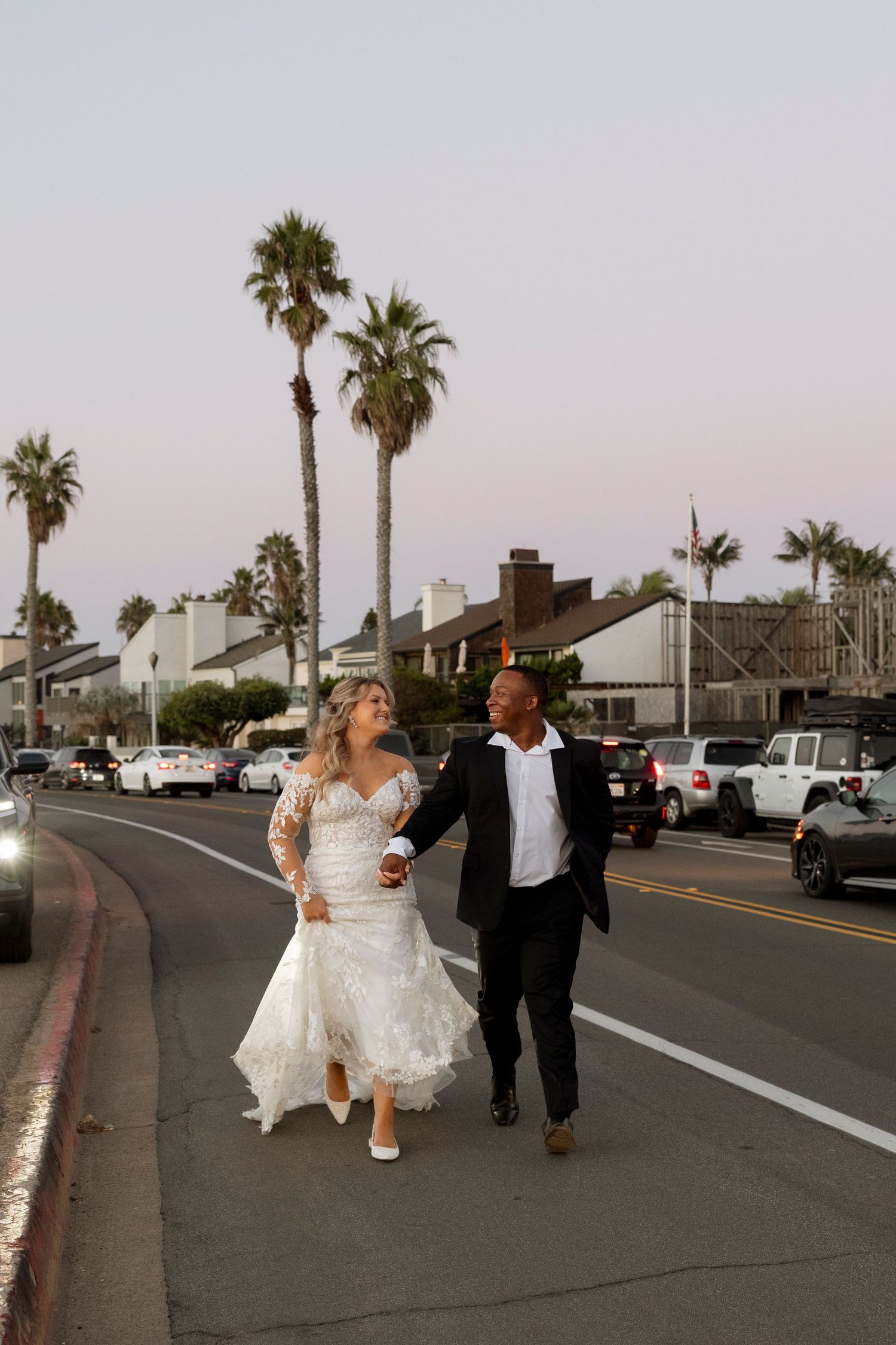 A couple in wedding attire walks down a palm tree-lined street at sunset.