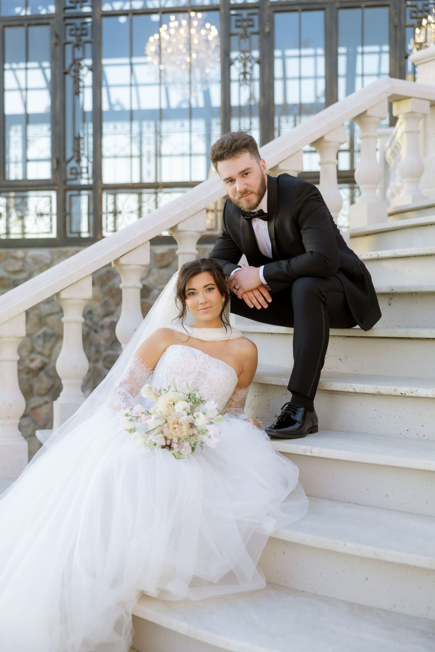 Bride in white gown holding bouquet sits on marble stairs with groom in black tuxedo at elegant wedding venue.