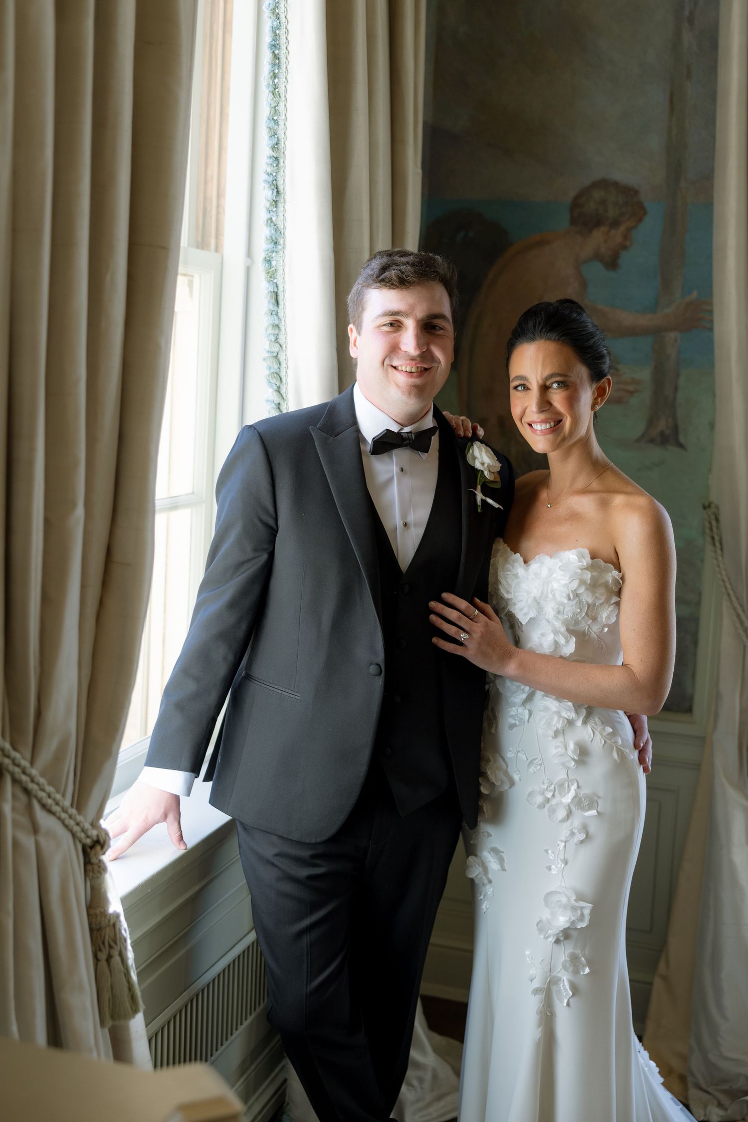 Smiling bride and groom posing by window in elegant venue with mural backdrop.