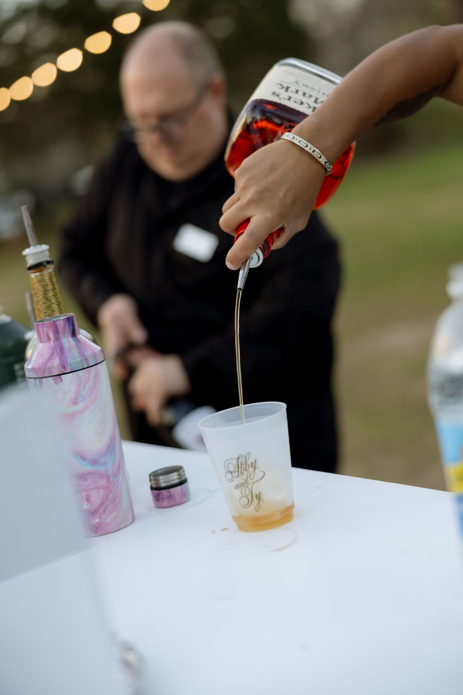 Bartender pours drink into personalized cup at outdoor wedding reception bar with string lights.