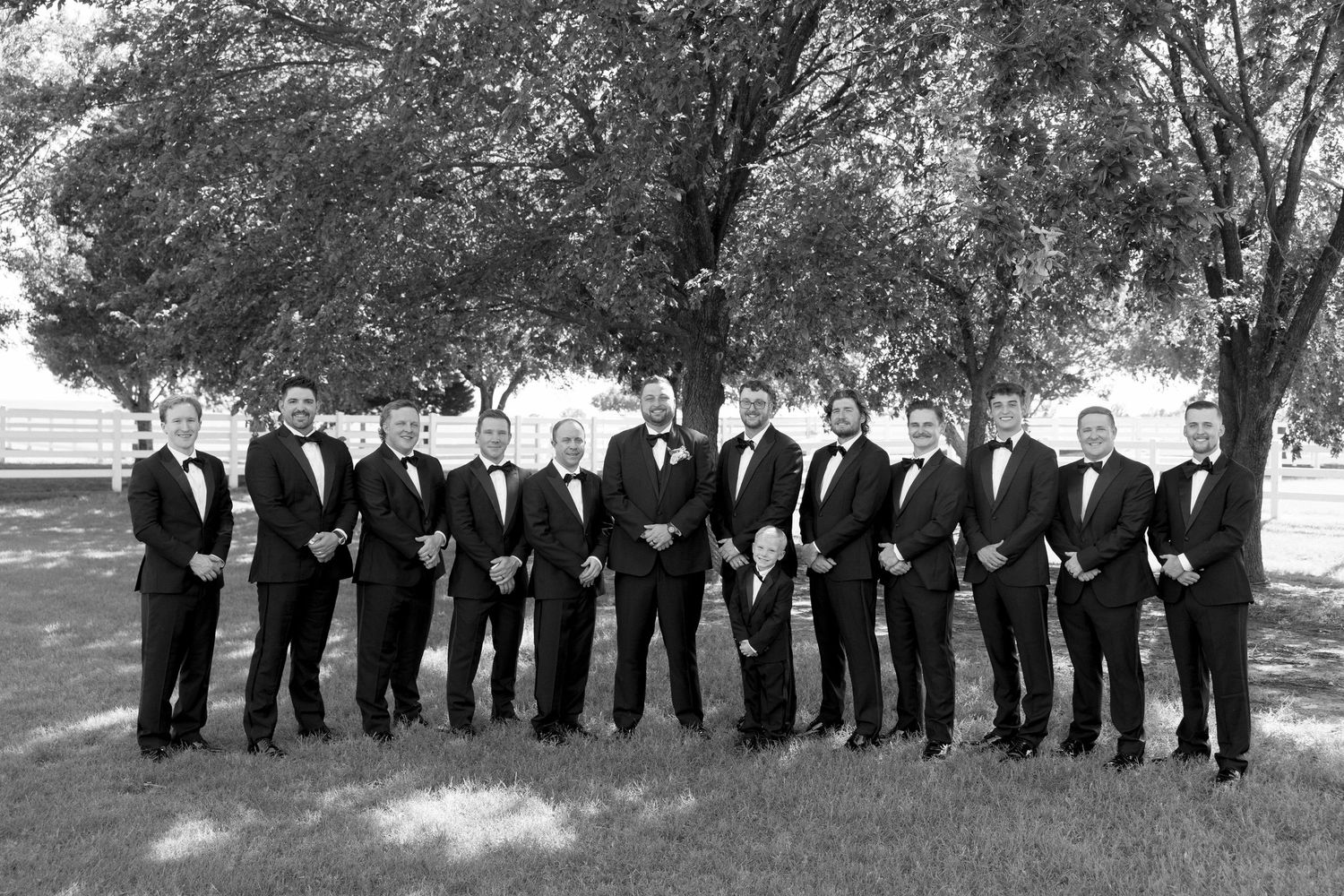 A formal group of groomsmen in black tuxedos standing in a line outdoors under trees.