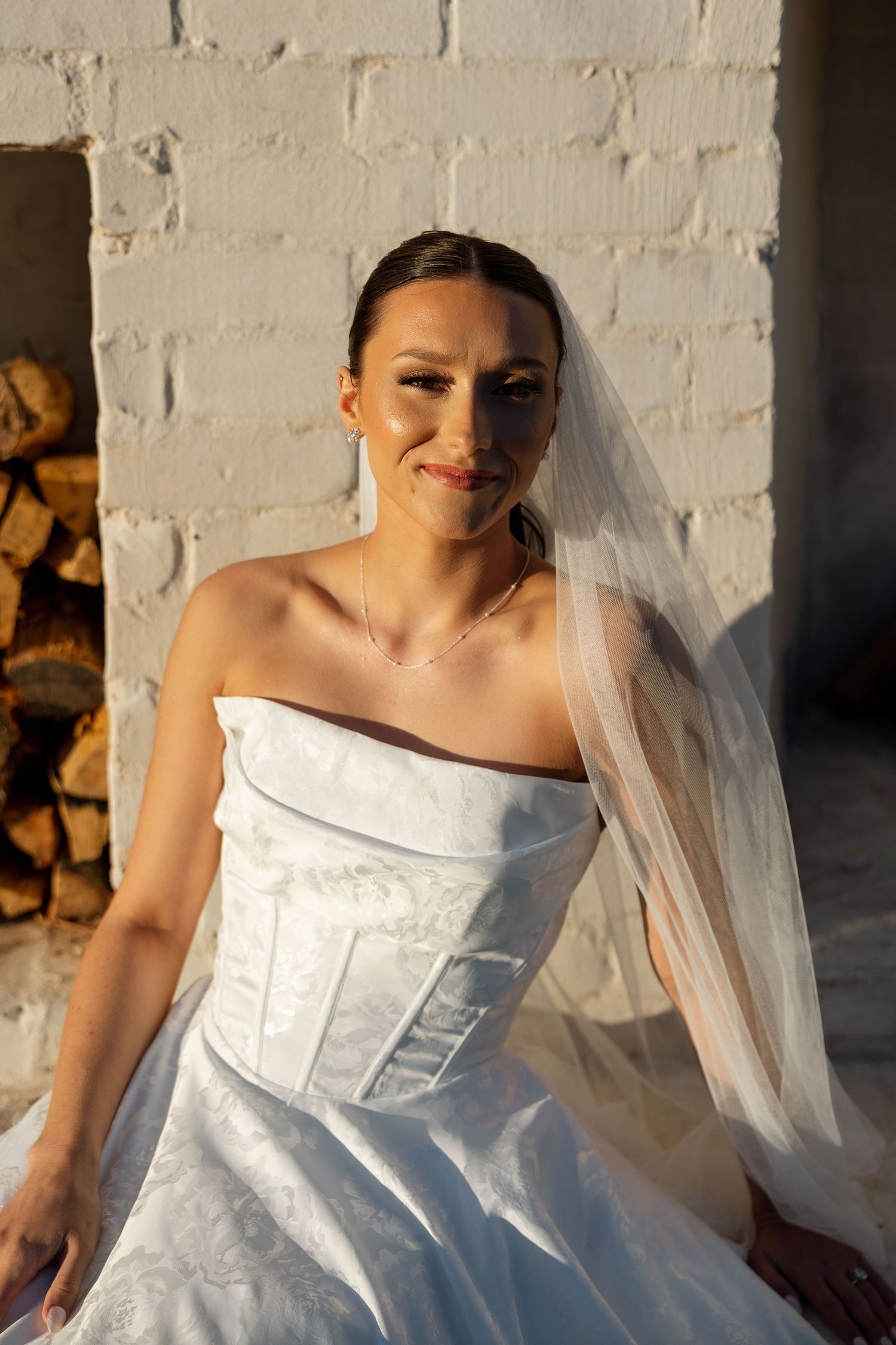 A bride in a strapless white wedding dress and veil smiles while posing against a sunlit white brick wall.