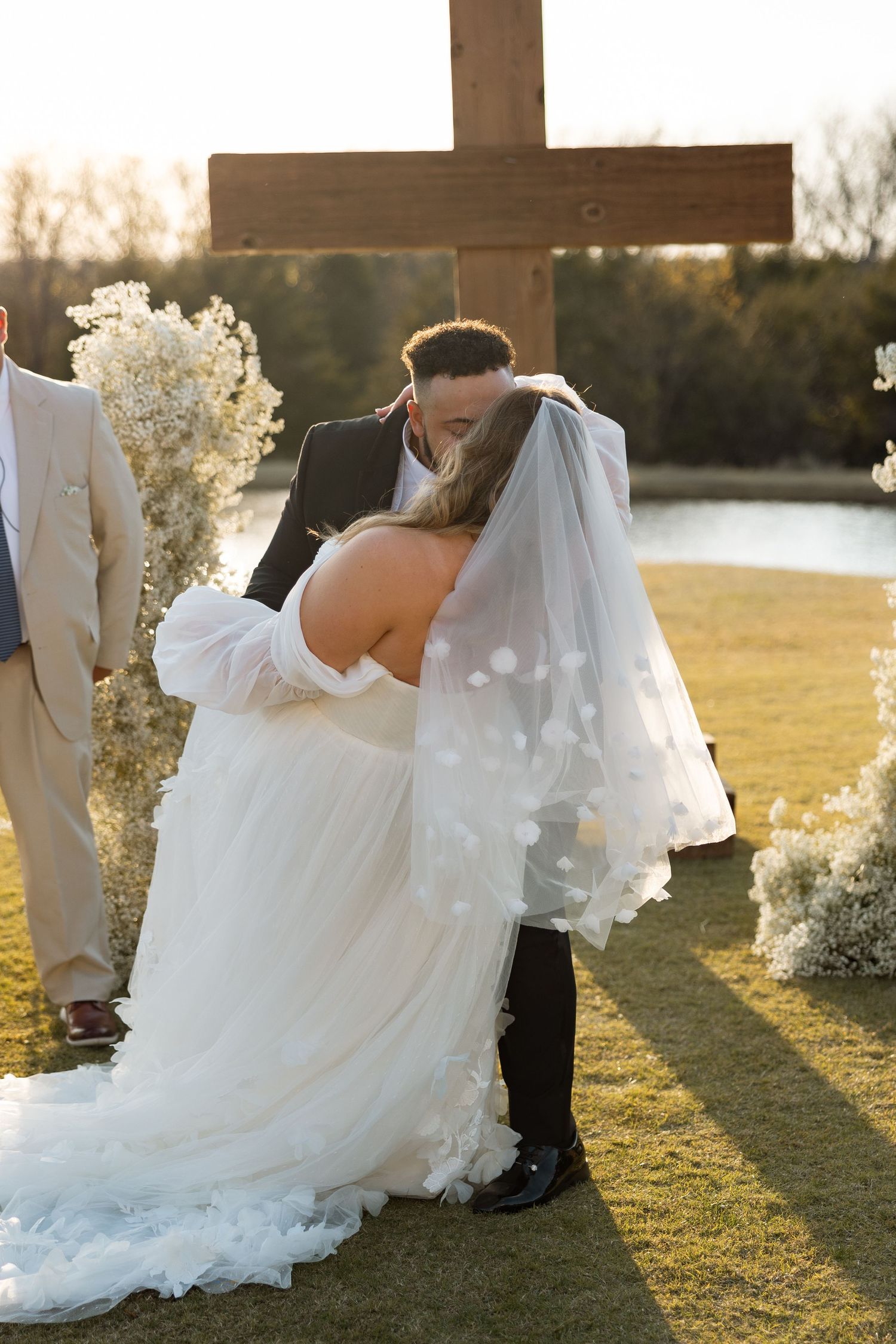 Romantic ceremony moment under wooden cross with lake and sunset in background.