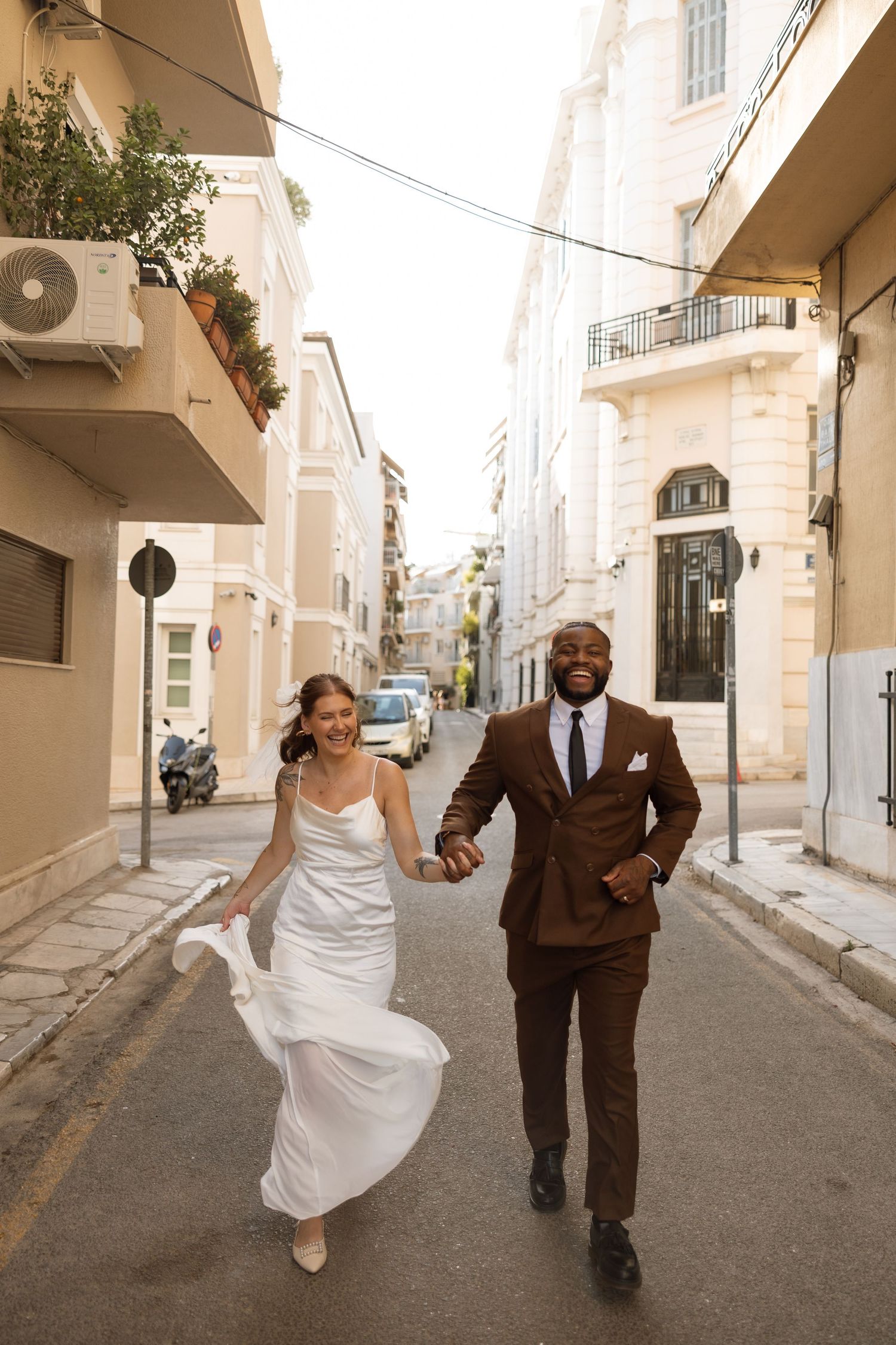 Newlyweds running joyfully down a charming European street with cream colored buildings and balconies.