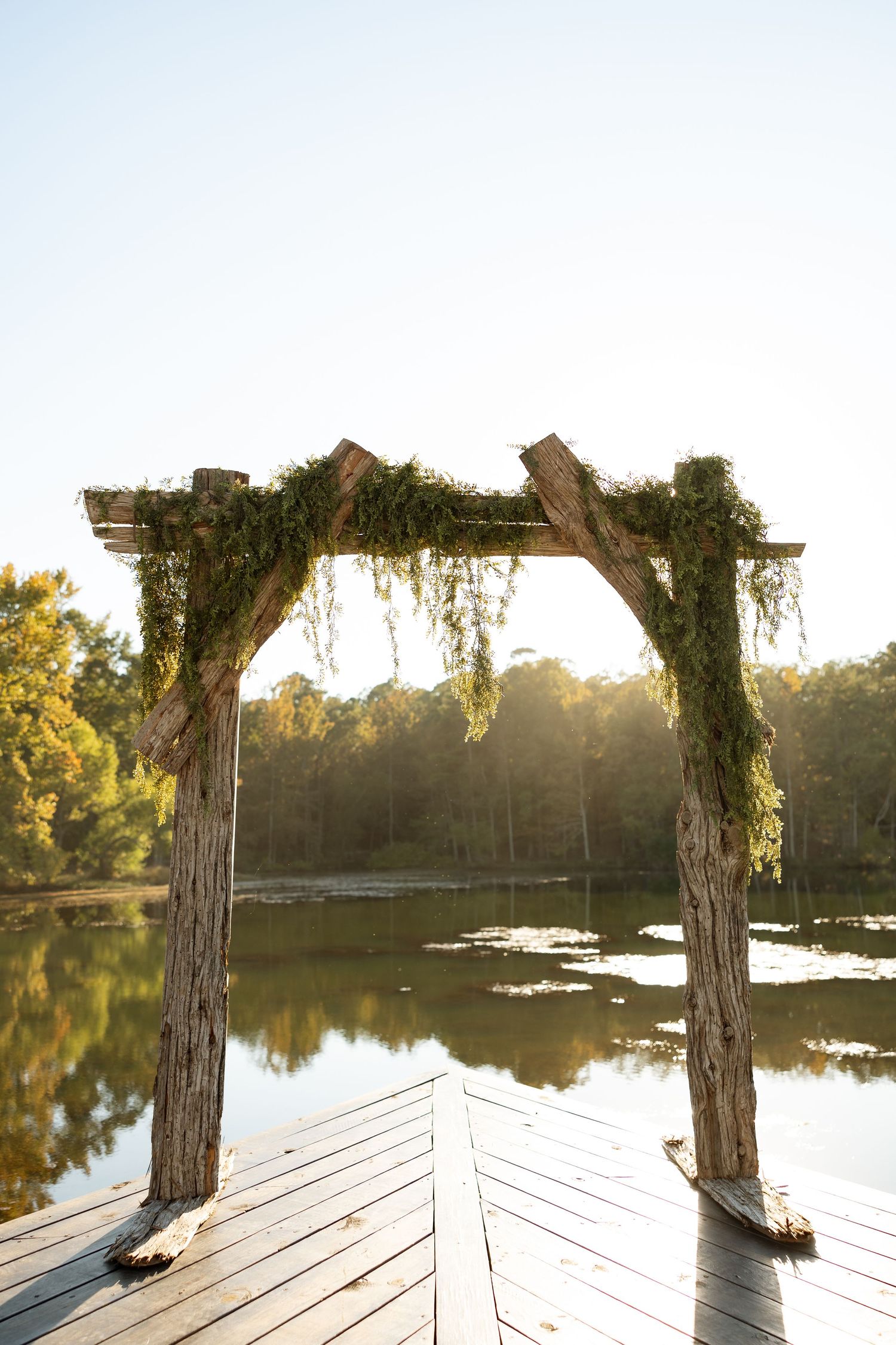 Rustic wooden wedding arch decorated with greenery overlooking a peaceful lake.