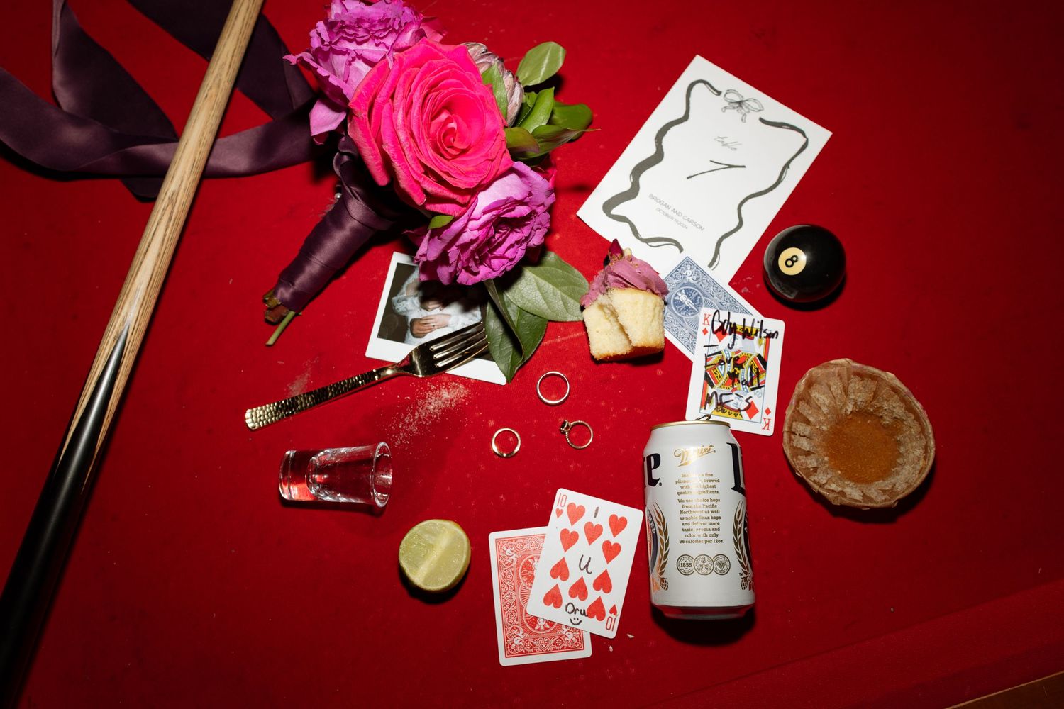 Wedding details flatlay with pink roses, playing cards and various decorative items on red background.
