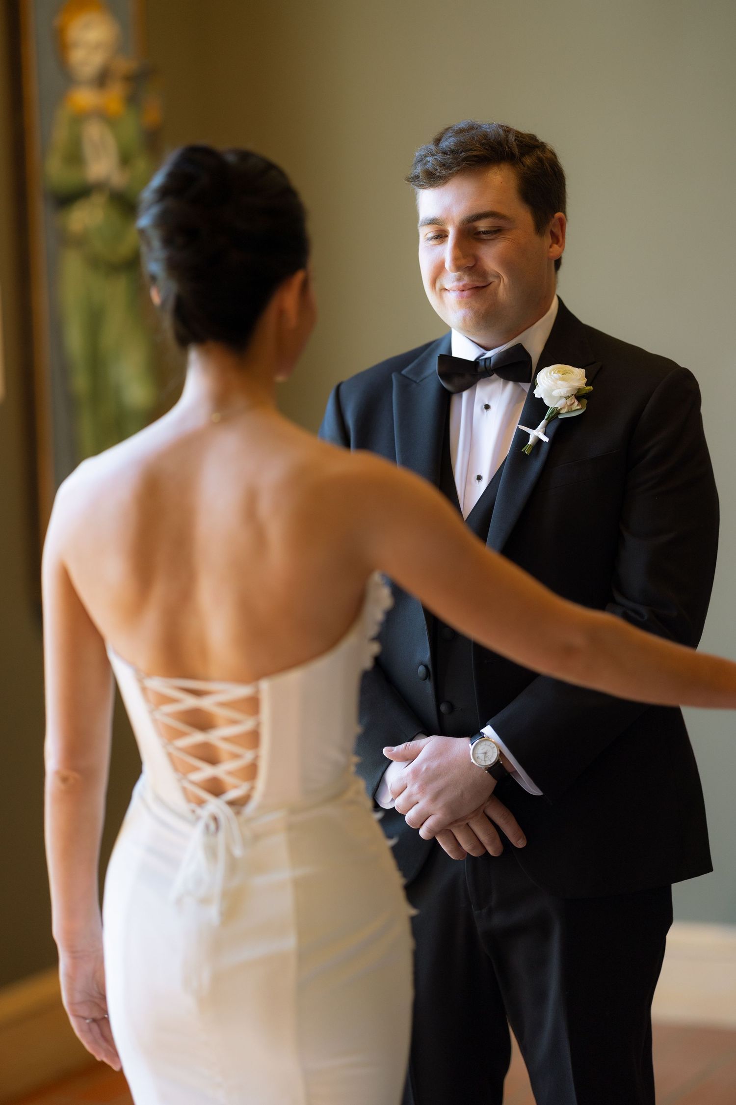 Groom in black tuxedo smiling at bride during emotional first look moment indoors.