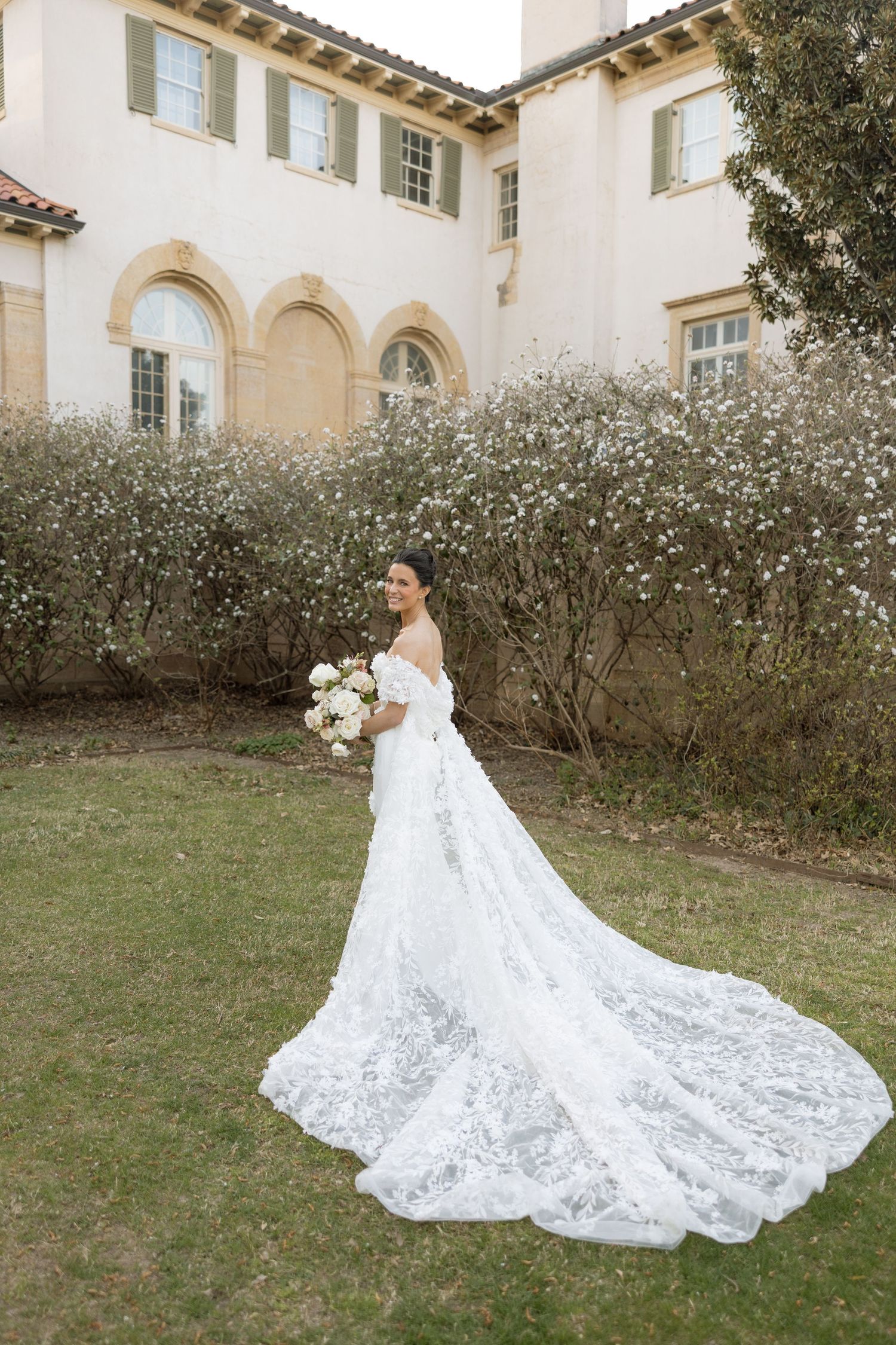 Bride in off-shoulder lace gown with long train poses in garden outside elegant estate.