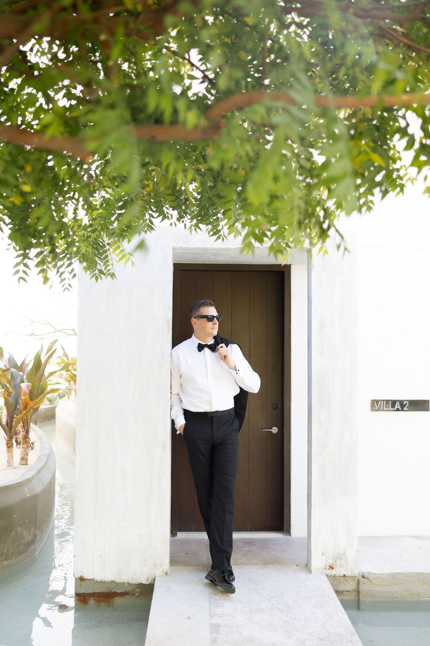 Groom in tuxedo with bow tie stands outside Villa 2 door beneath lush green tree canopy.