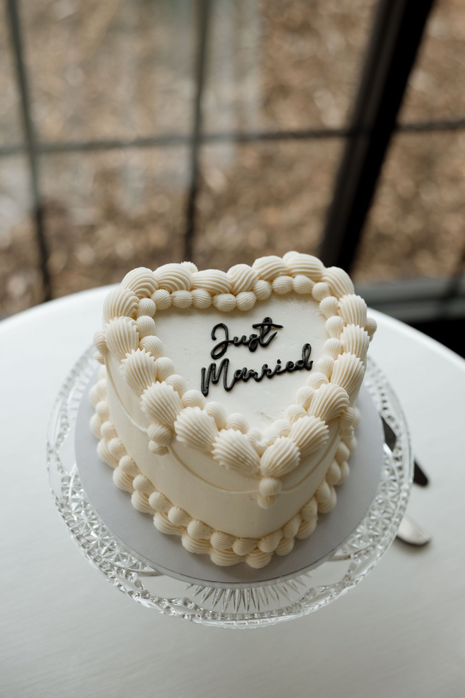Heart-shaped white wedding cake with shell piping and 'Just Married' text on crystal cake stand.
