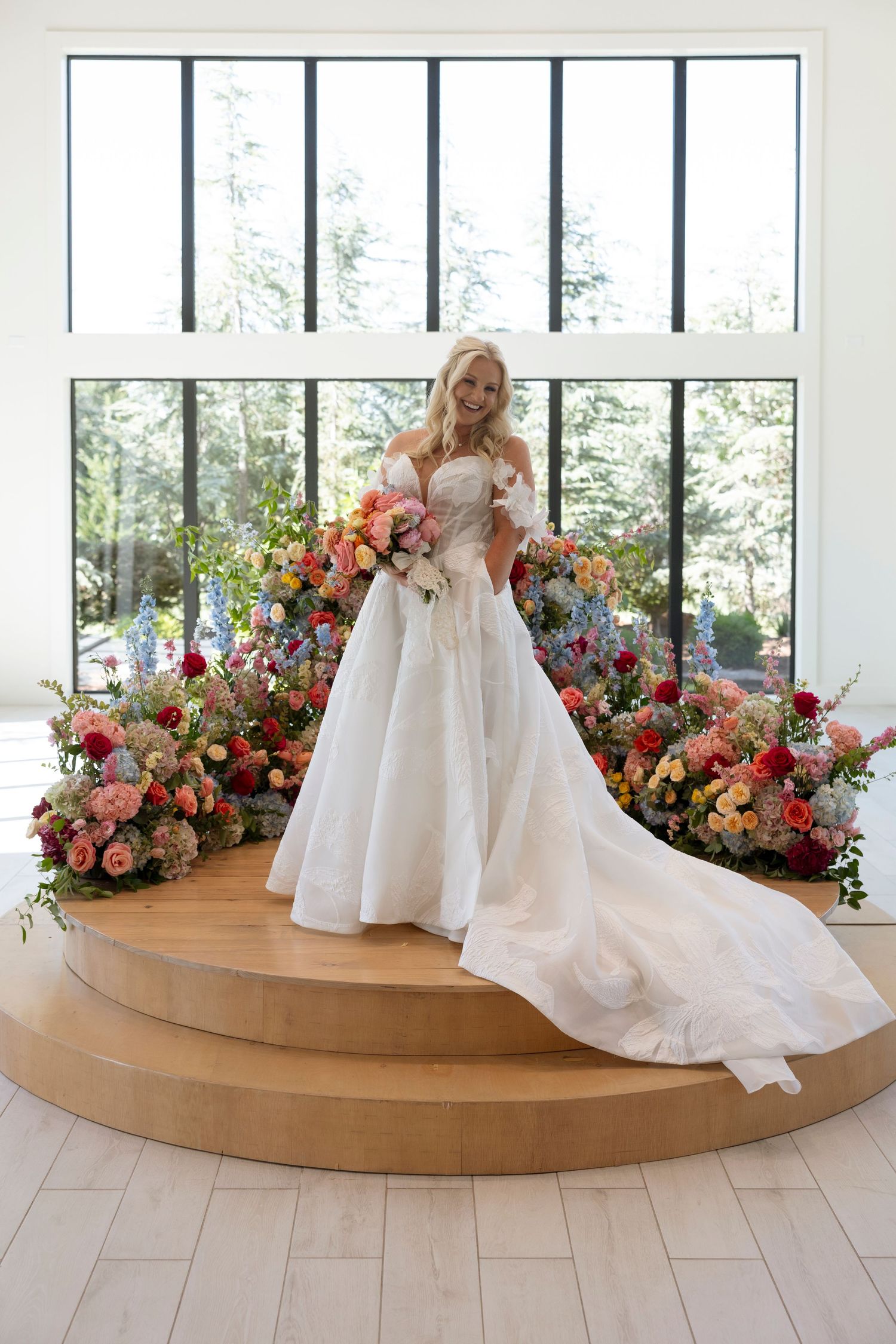 Bride in flowing white gown poses on curved wooden platform surrounded by vibrant floral arrangements.